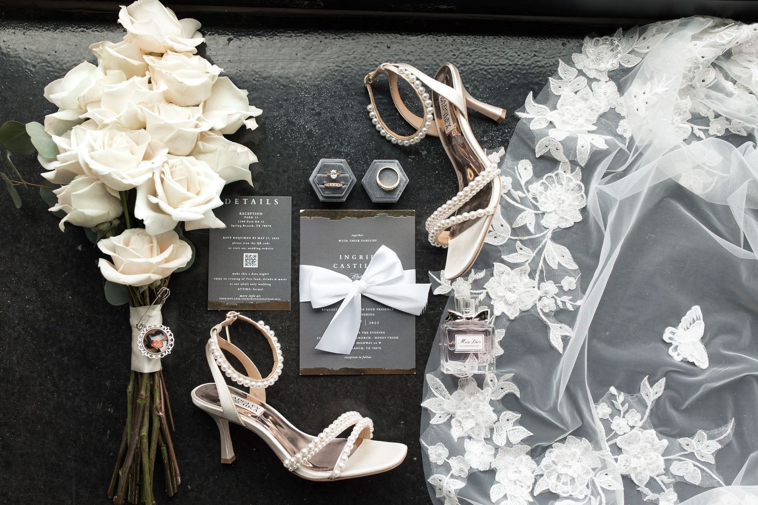 Flat lay wedding detail photo with bridal bouquet, perfume bottle, wedding invitation suite, heels, and lace veil on dark background.