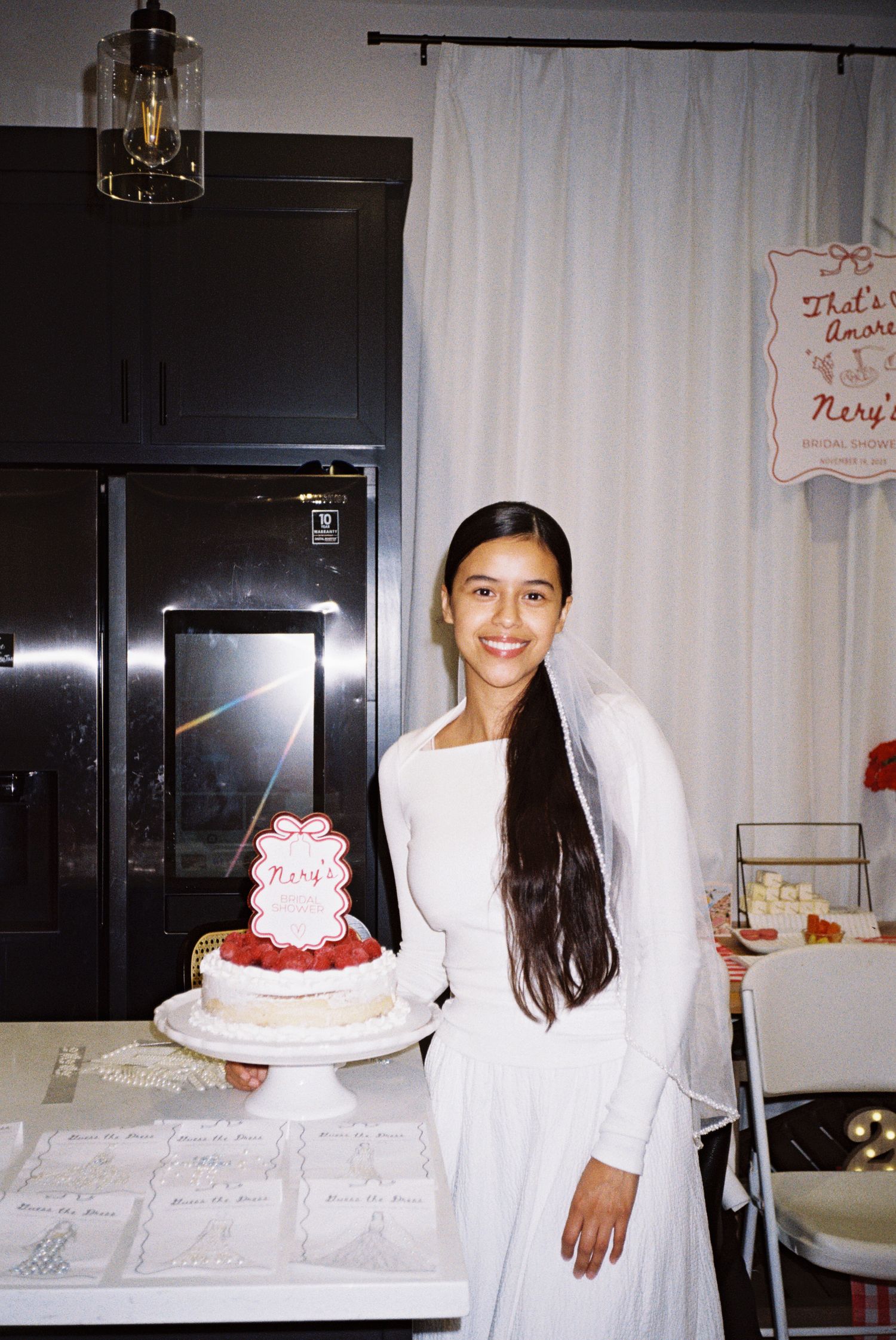 Bride in white dress smiling indoors, holding tiered wedding cake in modern kitchen setting.