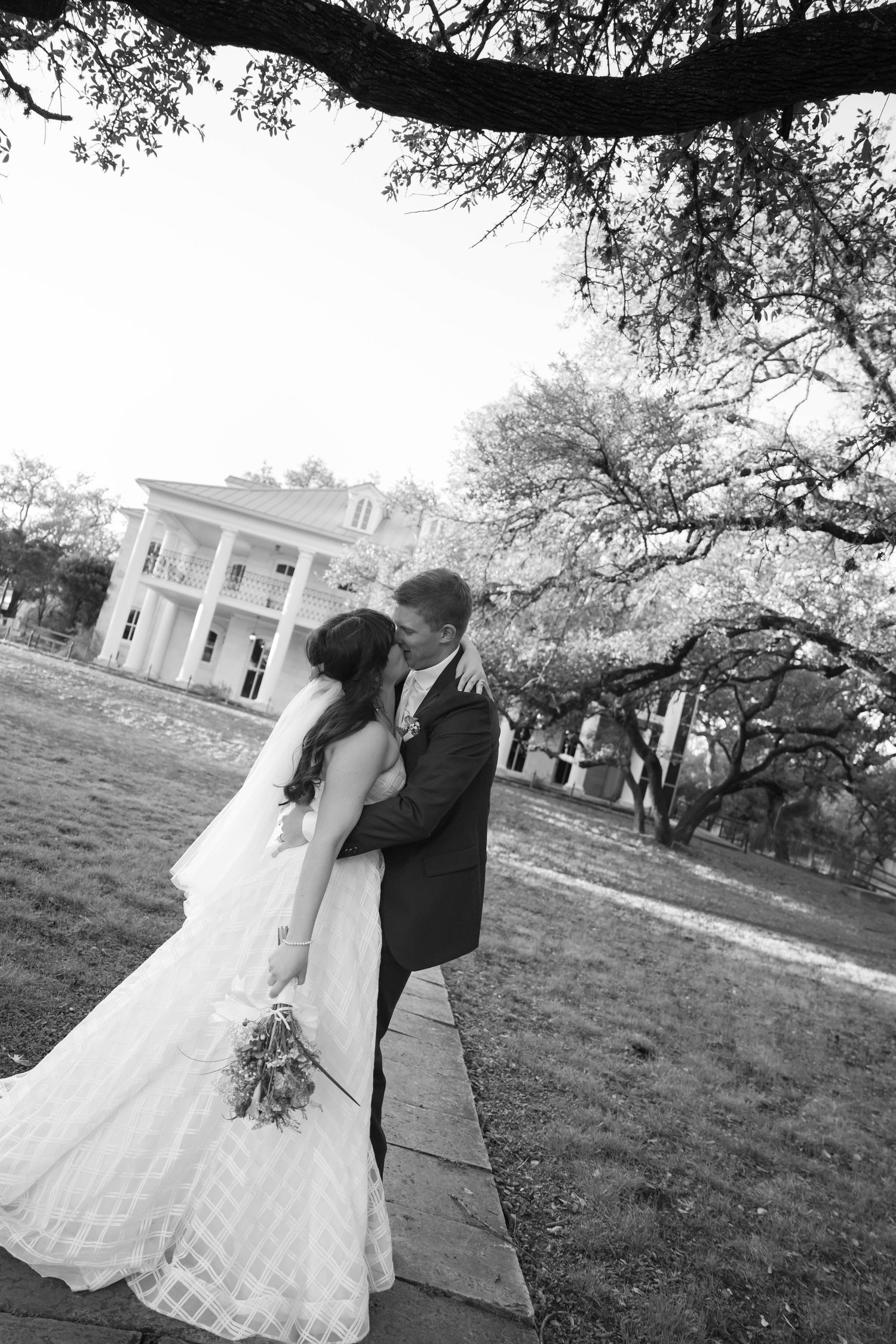 Black and white outdoor wedding portrait of bride and groom embracing under large oak tree with historic mansion in the background.