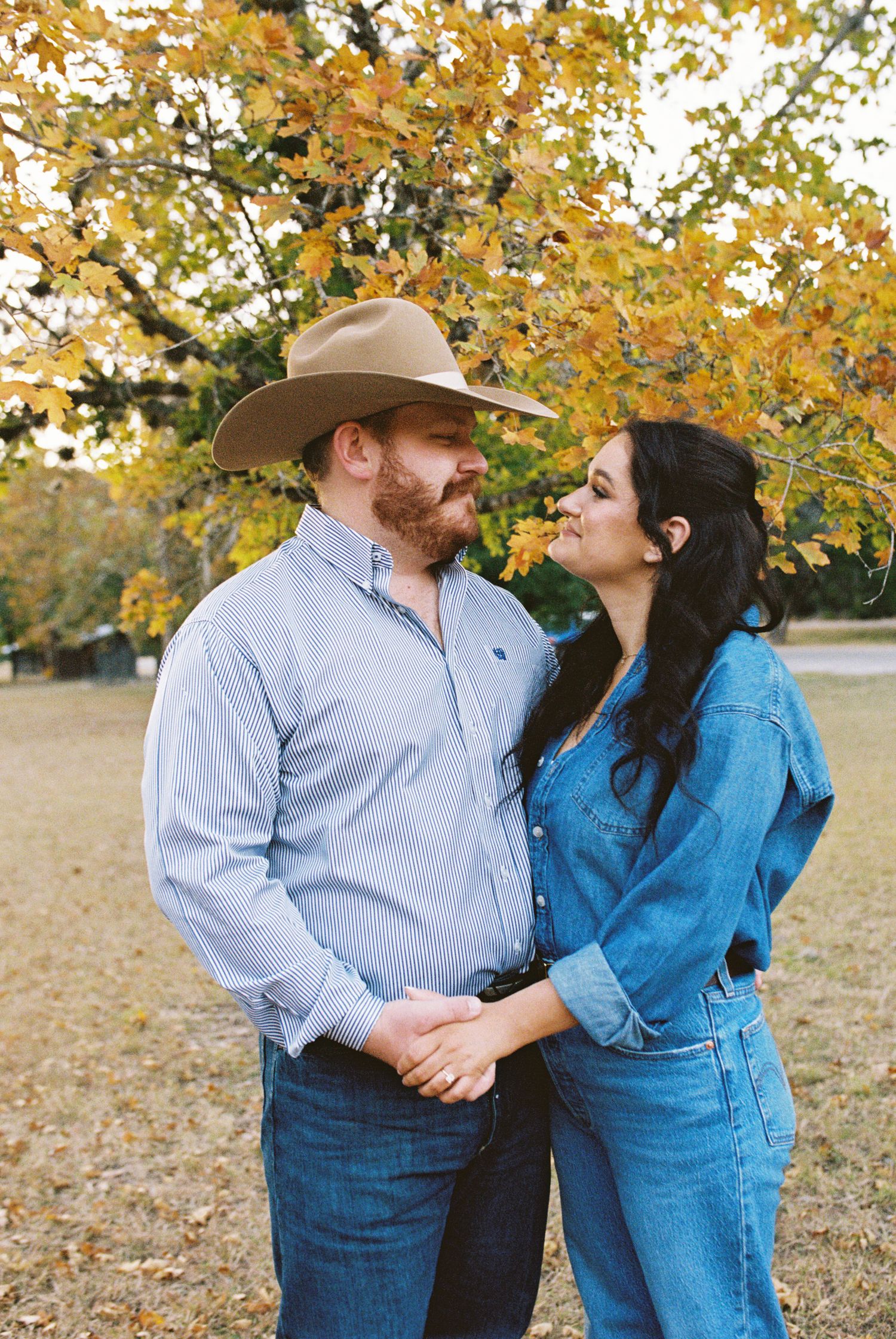 Couple standing close together under golden autumn trees, man wearing a cowboy hat and light blue shirt, woman in denim outfit smiling at ea