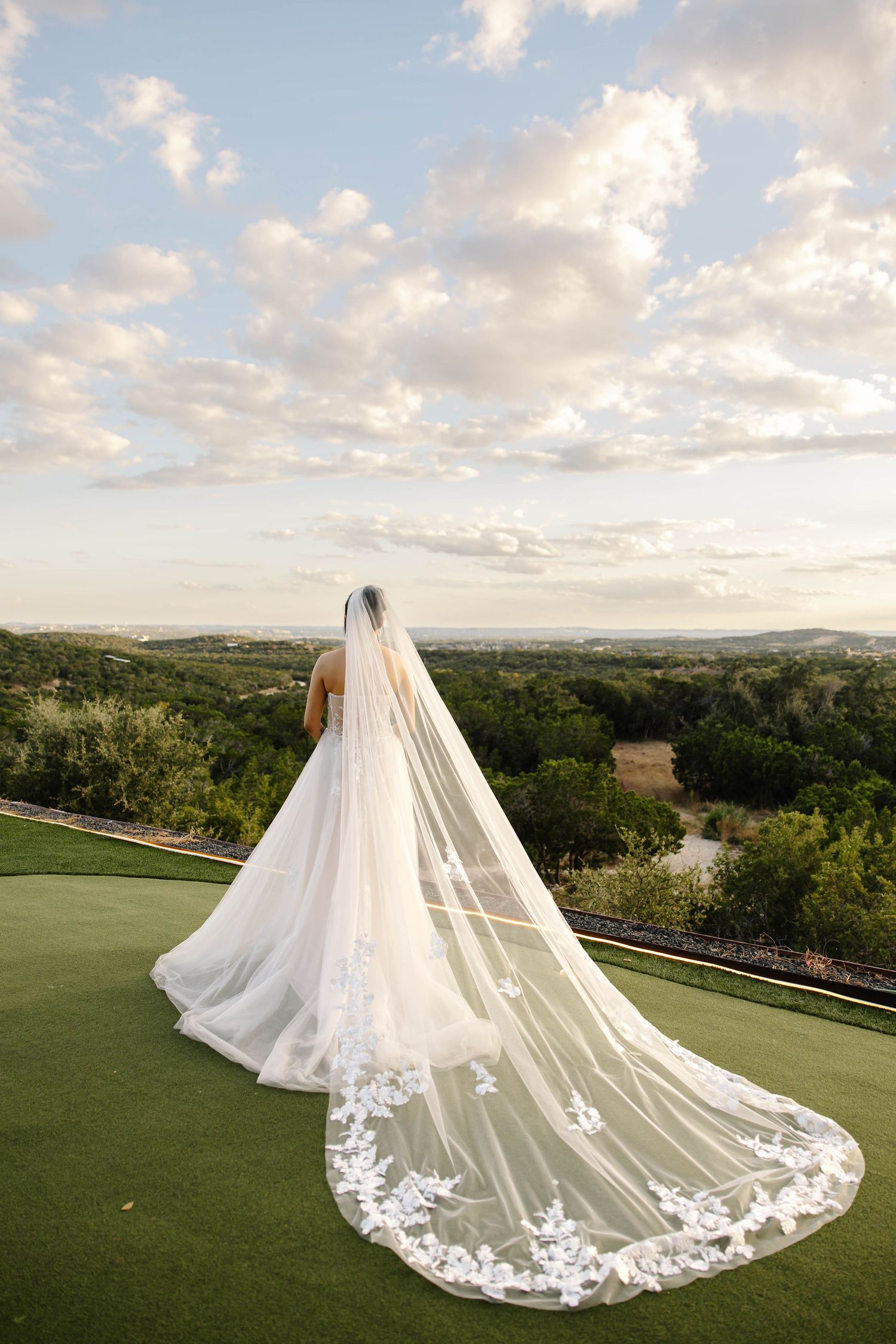 Bride standing on a hill overlooking scenic landscape with long lace cathedral veil flowing behind her at sunset.