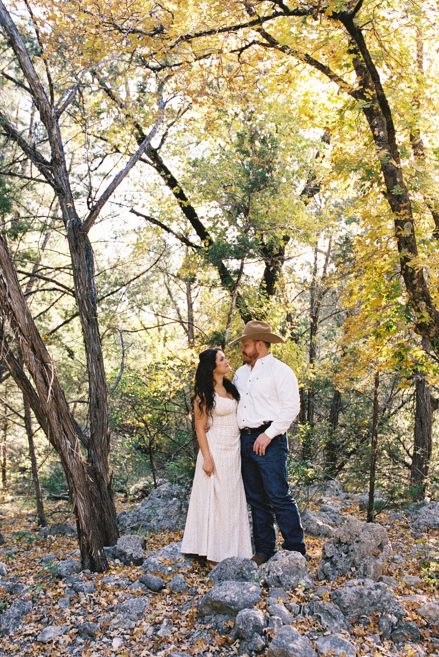 Couple standing among trees in fall foliage, embracing in soft sunlight, romantic woodland engagement portrait.
