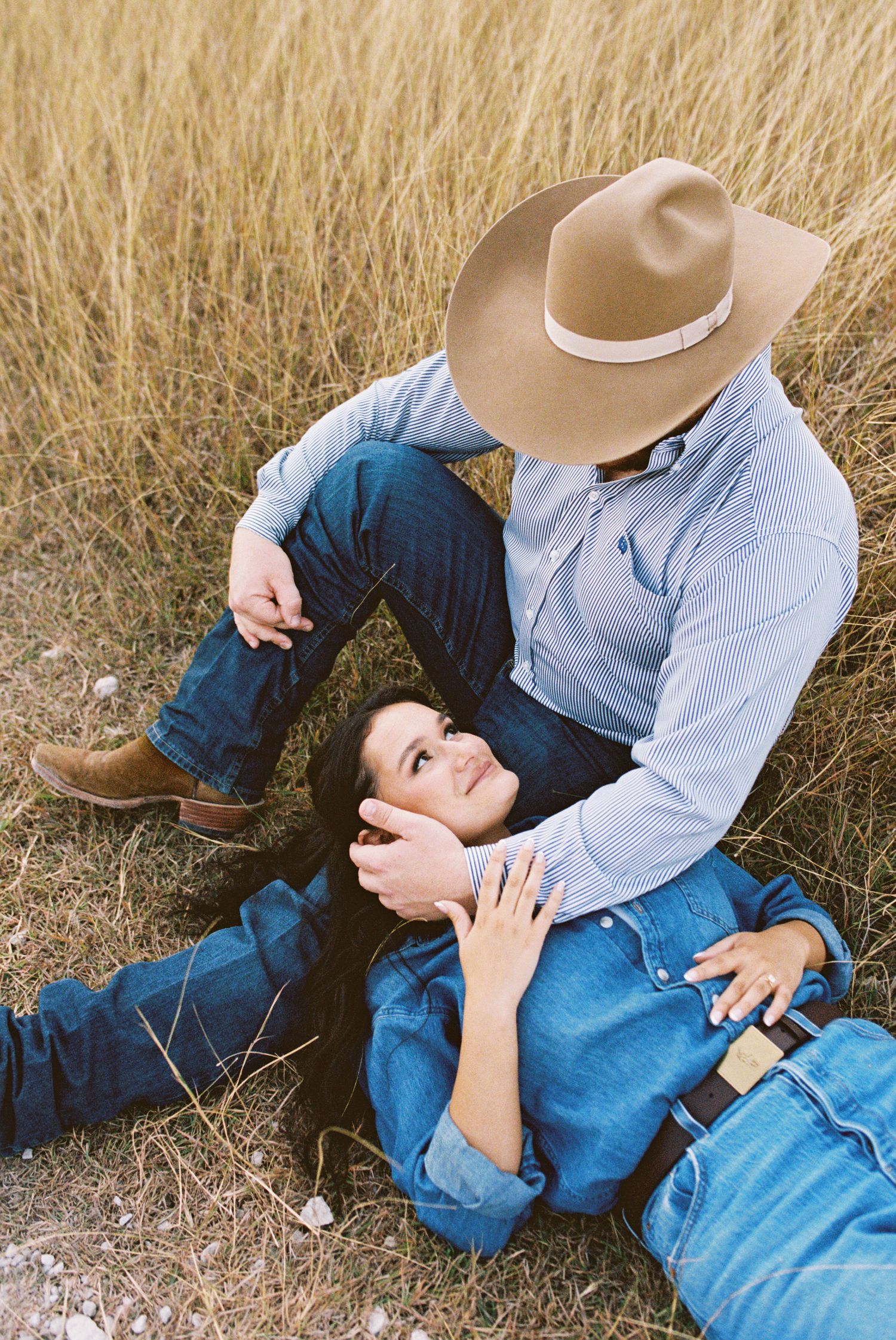 Close-up of couple sitting on wheat grass, hands intertwined showing engagement ring, casual denim outfits in rustic setting.