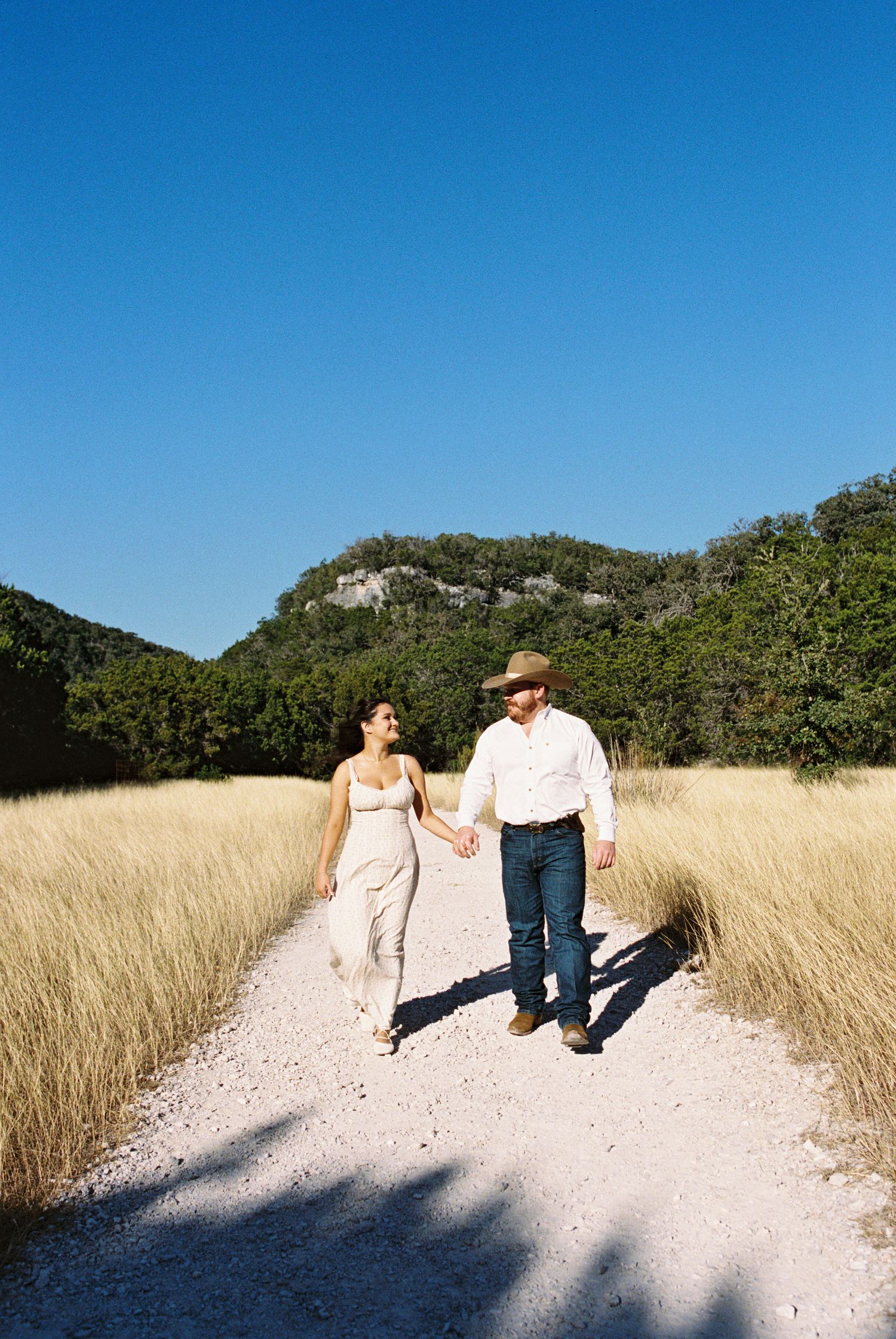 Couple walking hand in hand down dirt path through tall grass, blue sky and scenic rural landscape.