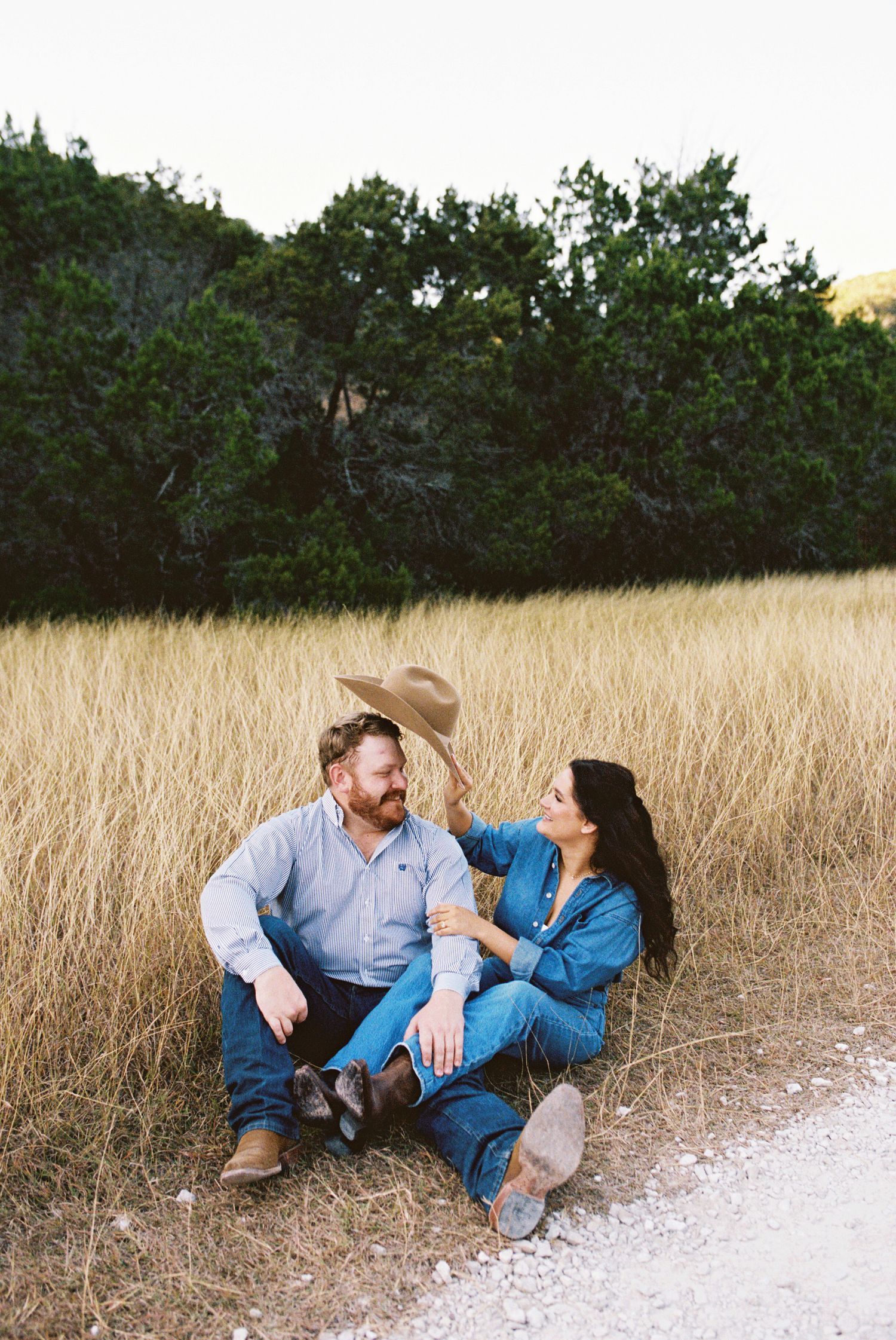 Couple lounging in dry grass field, smiling and relaxed, countryside engagement session with natural tones.