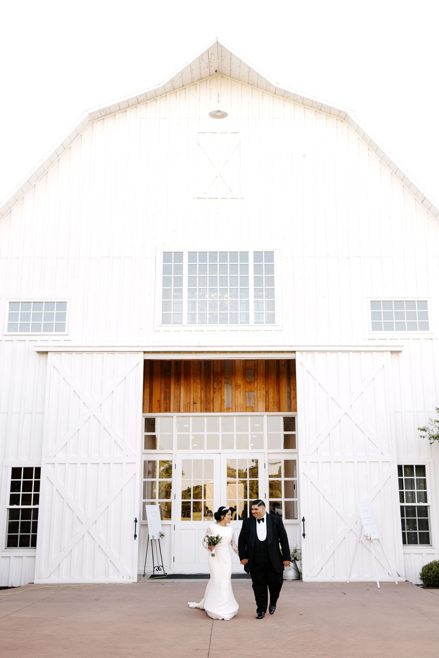 Bride and groom walking out of a large white barn wedding venue with tall doors and rustic modern architecture.