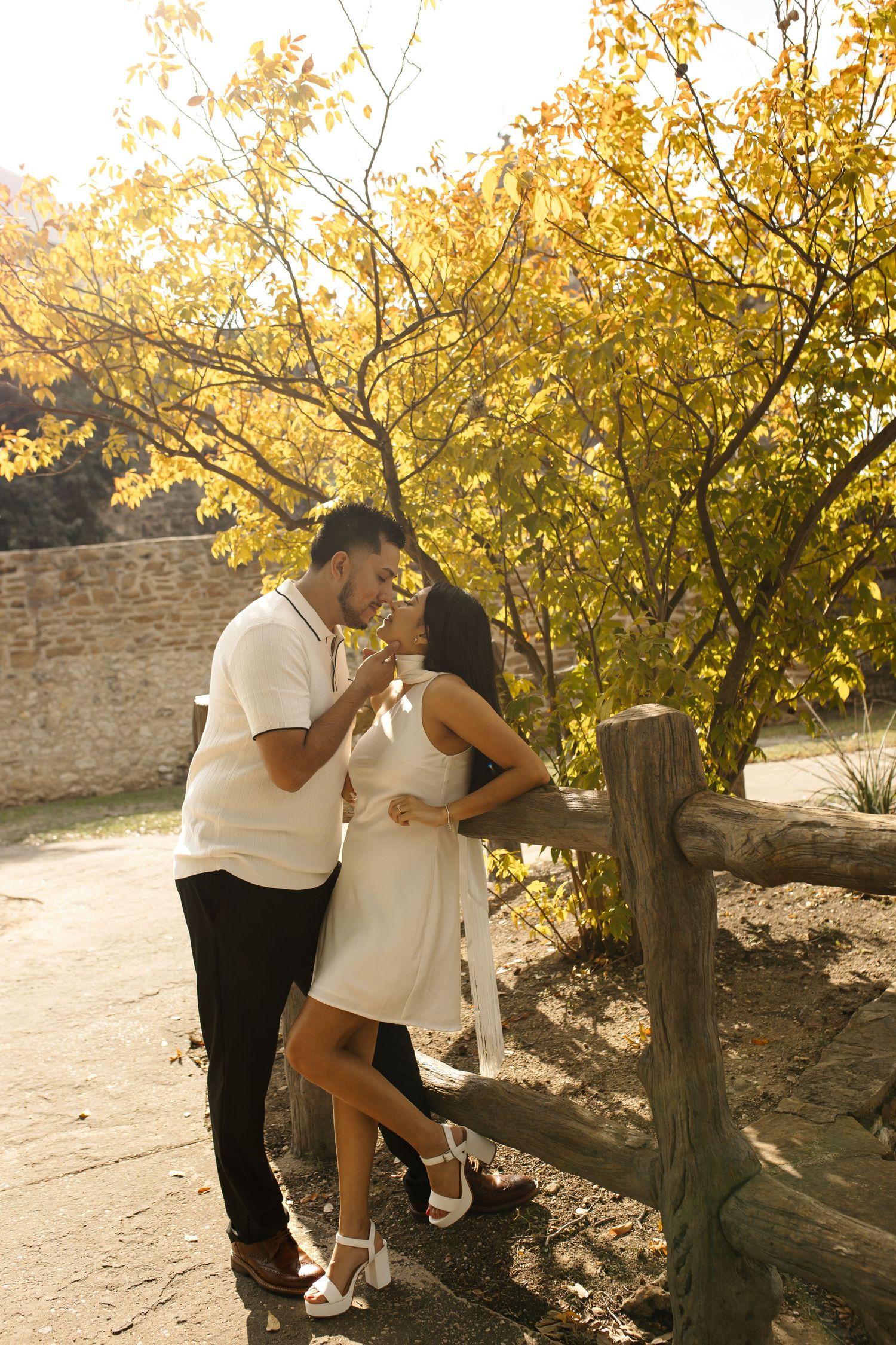 Romantic couple kissing by rustic wooden fence under golden autumn trees, warm sunlight, fall park setting, intimate outdoor moment.