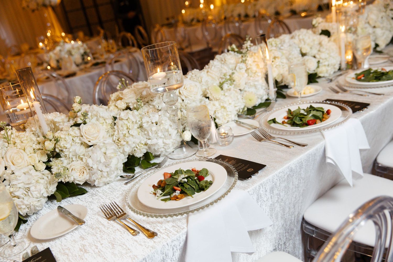 Wedding reception tablescape with white floral centerpieces, gold flatware, plated salads, and candles on an elegant long banquet table.