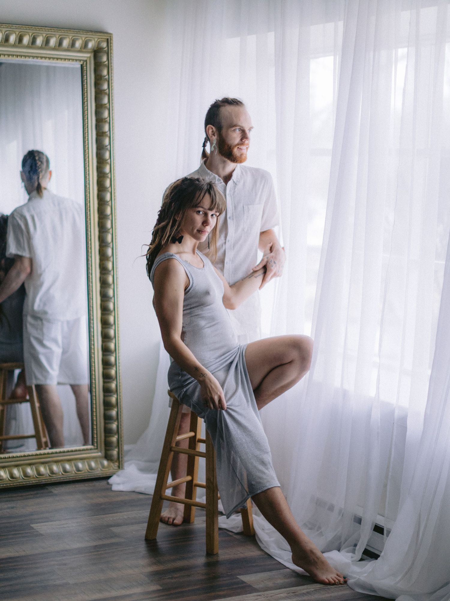 A couple poses elegantly near sheer curtains beside a gold-framed mirror, woman seated on stool in gray dress.