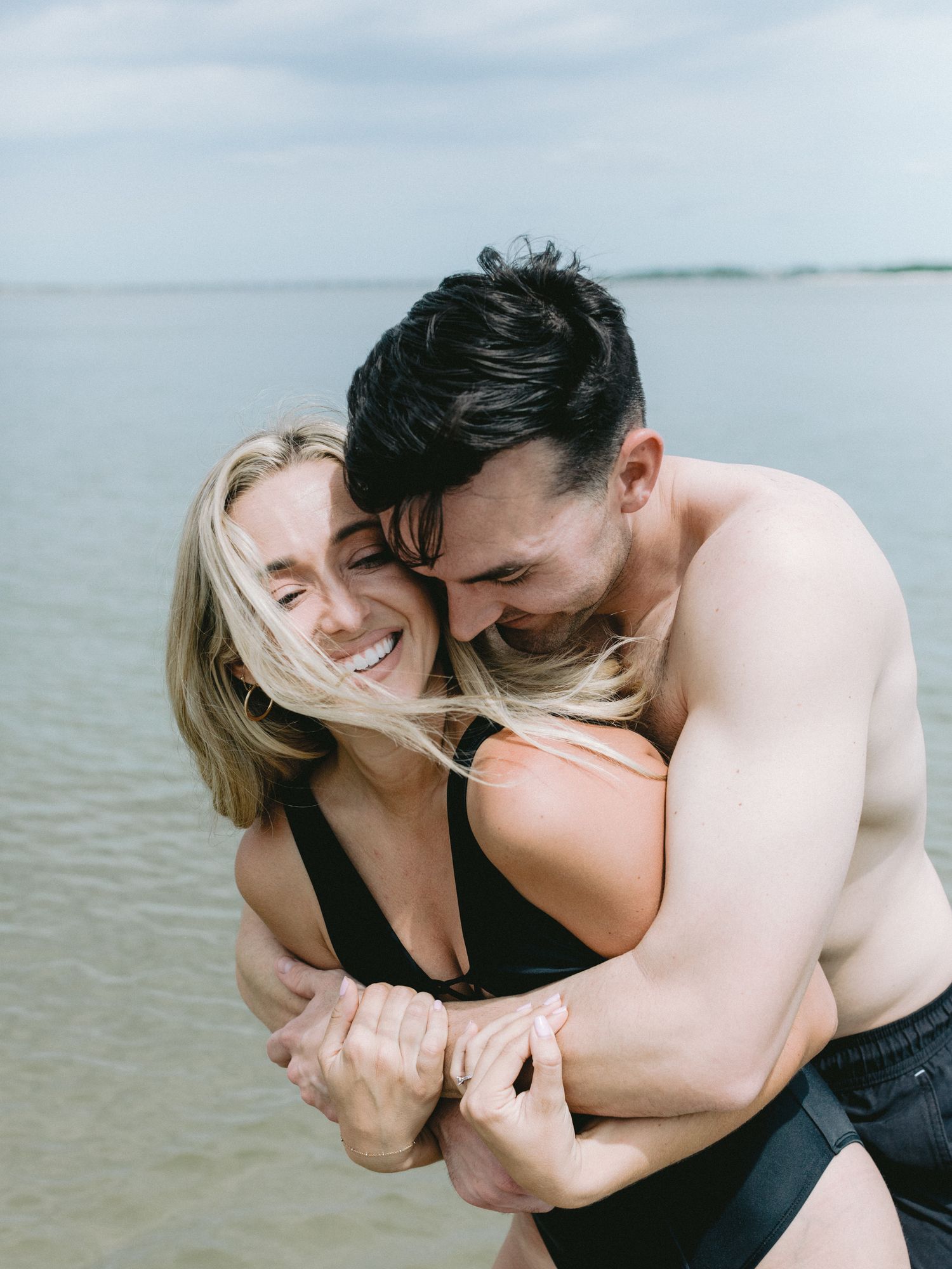 Happy couple embracing and laughing together in black swimwear at a calm waterfront.
