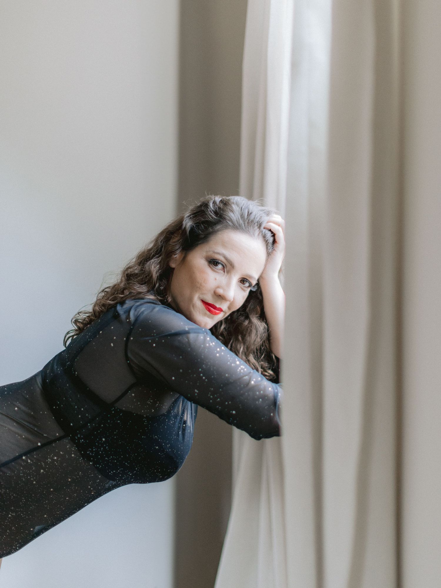 Brunette woman in a sparkling dark bodysuit leaning against a curtain with red lips.