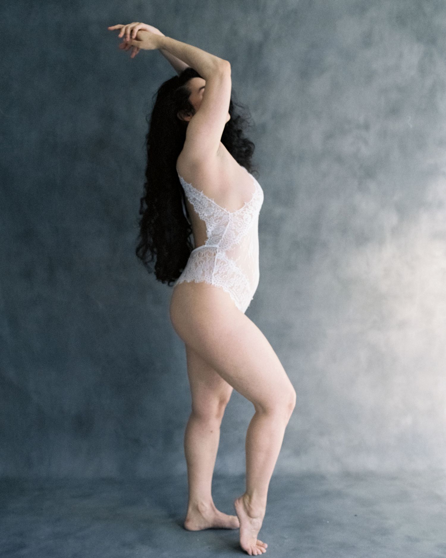 Woman in white lace bodysuit poses gracefully against gray backdrop, showing elegant side profile with arms raised.