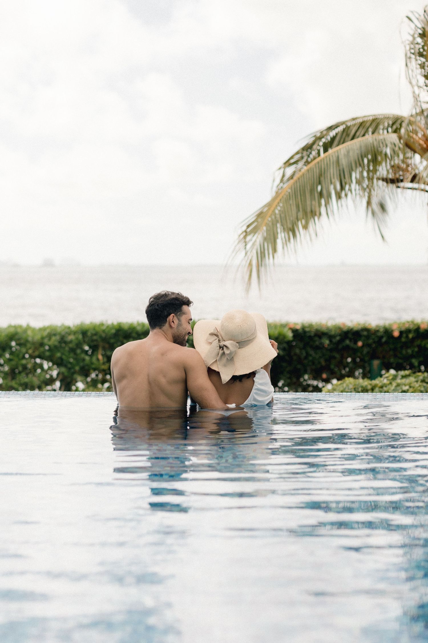 Romantic couple kissing in infinity pool overlooking ocean, woman wearing wide-brim sun hat.