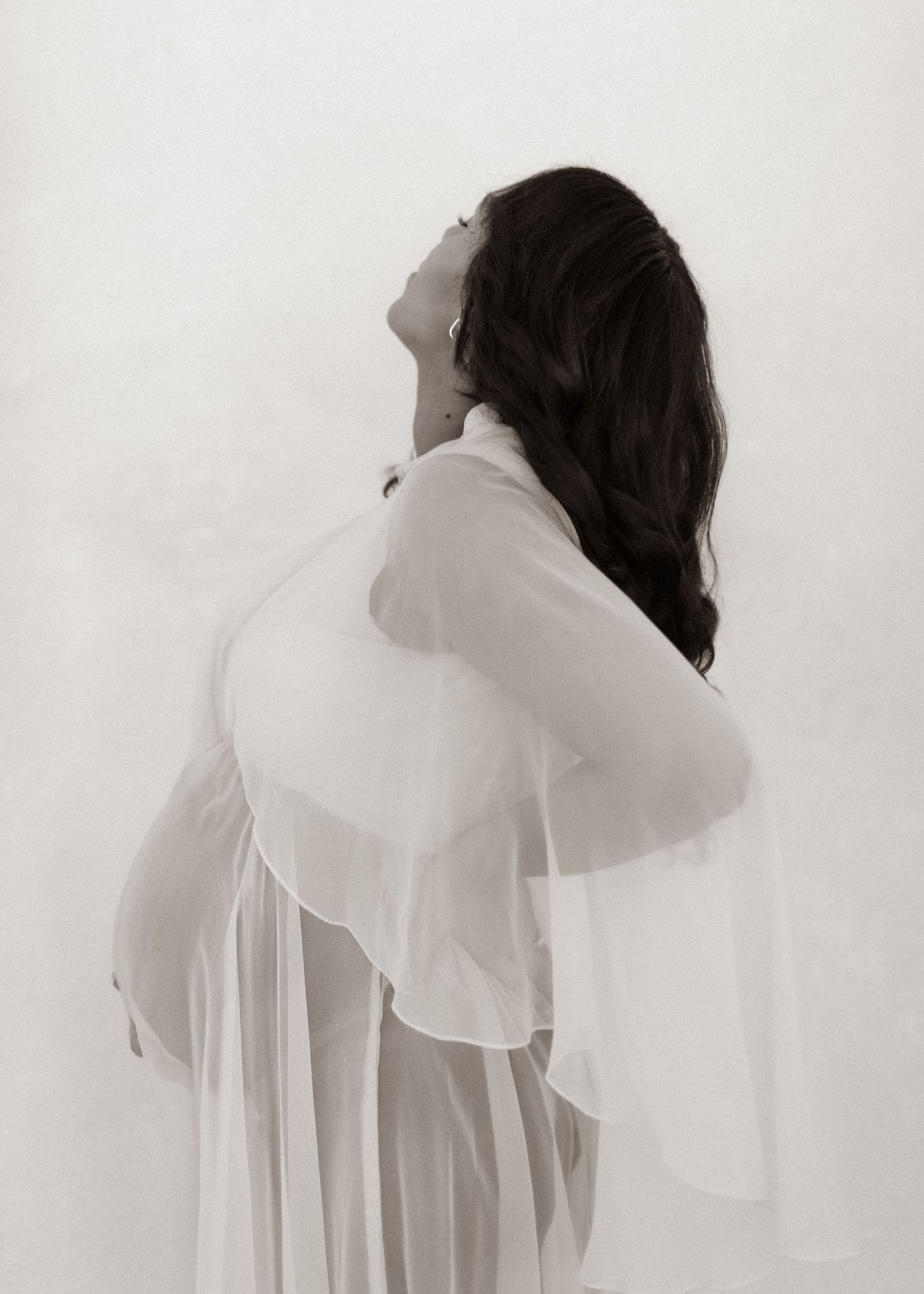 Pregnant woman in sheer white gown poses sideways, head tilted back, long dark hair flowing, in elegant maternity portrait.