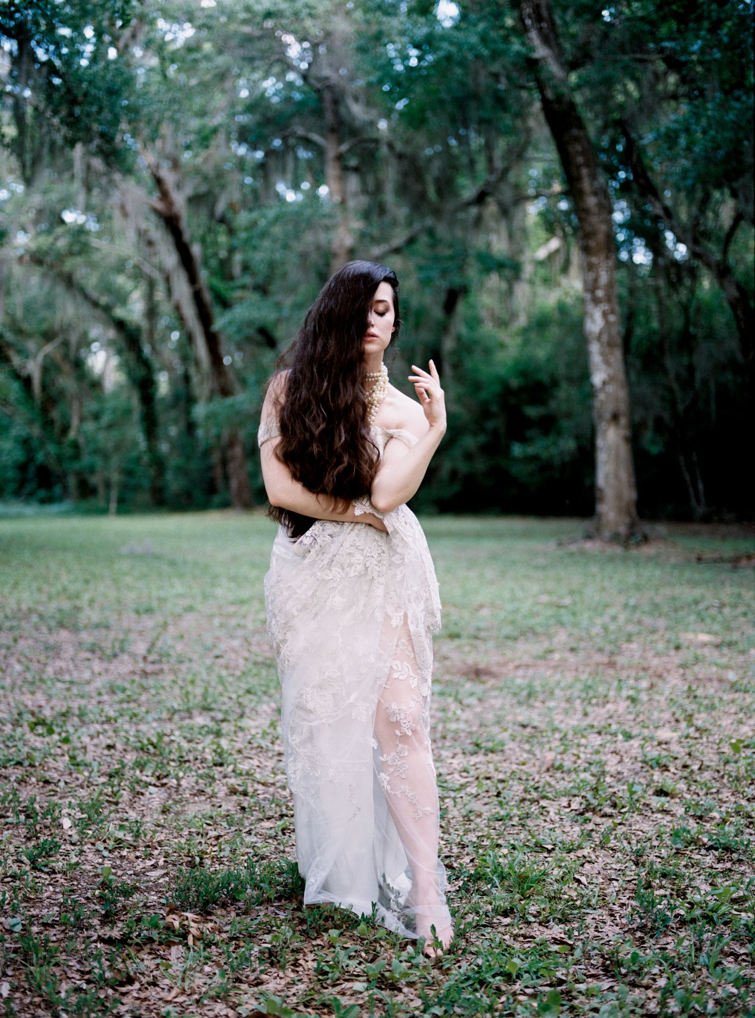 Woman in sheer floral gown standing in a lush green outdoor setting with long flowing hair.