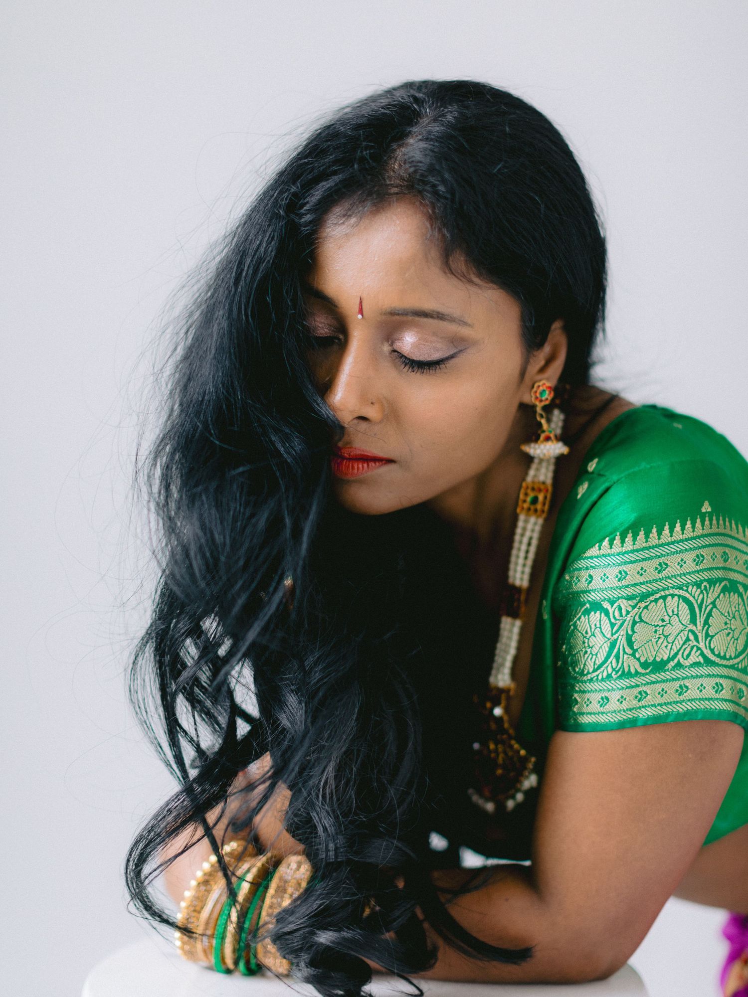 Indian woman in green silk saree with gold jewelry and long black hair poses gracefully in a studio portrait.