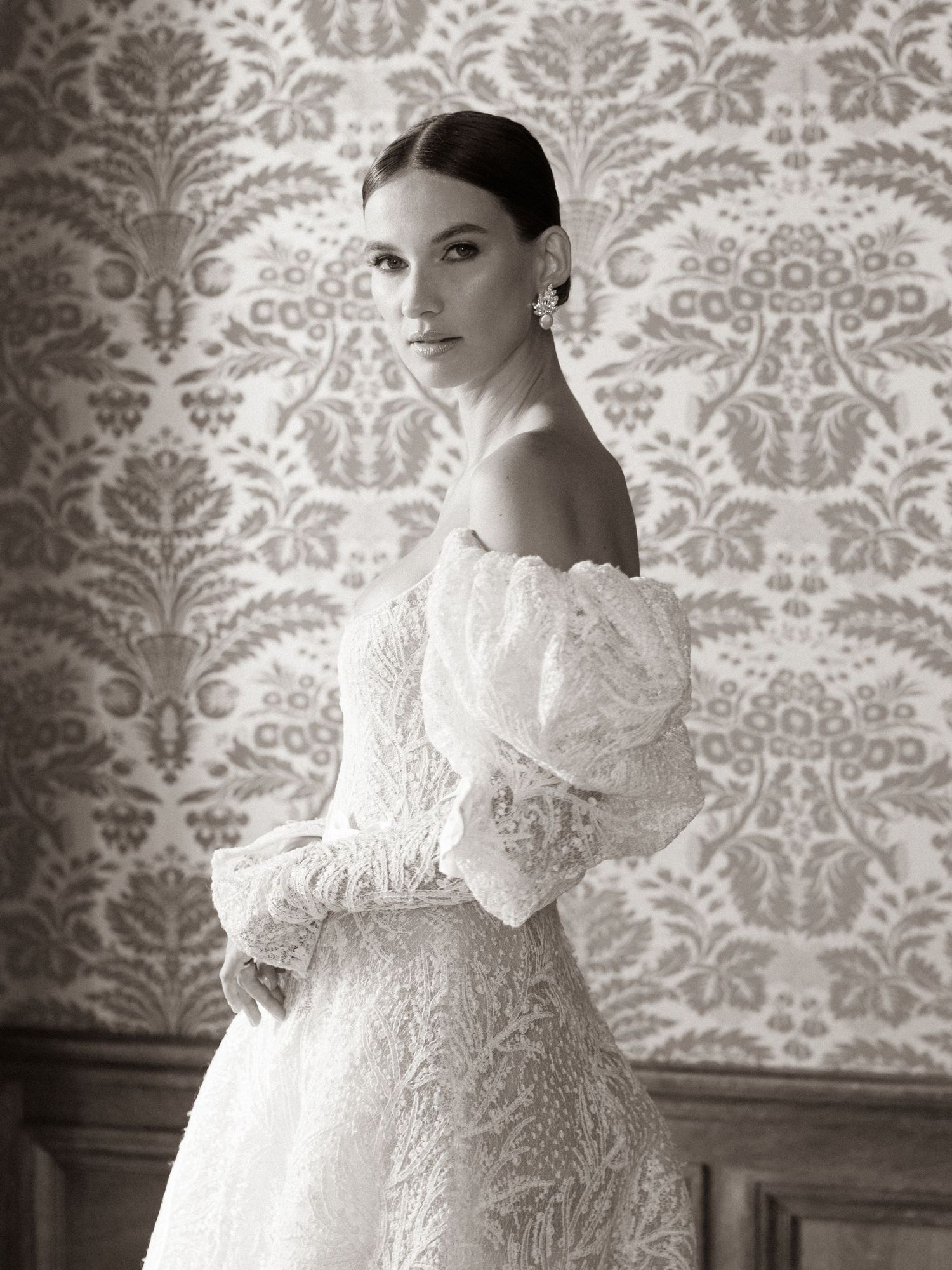 Black and white bridal portrait of woman in off-shoulder lace wedding gown before ornate wallpaper.