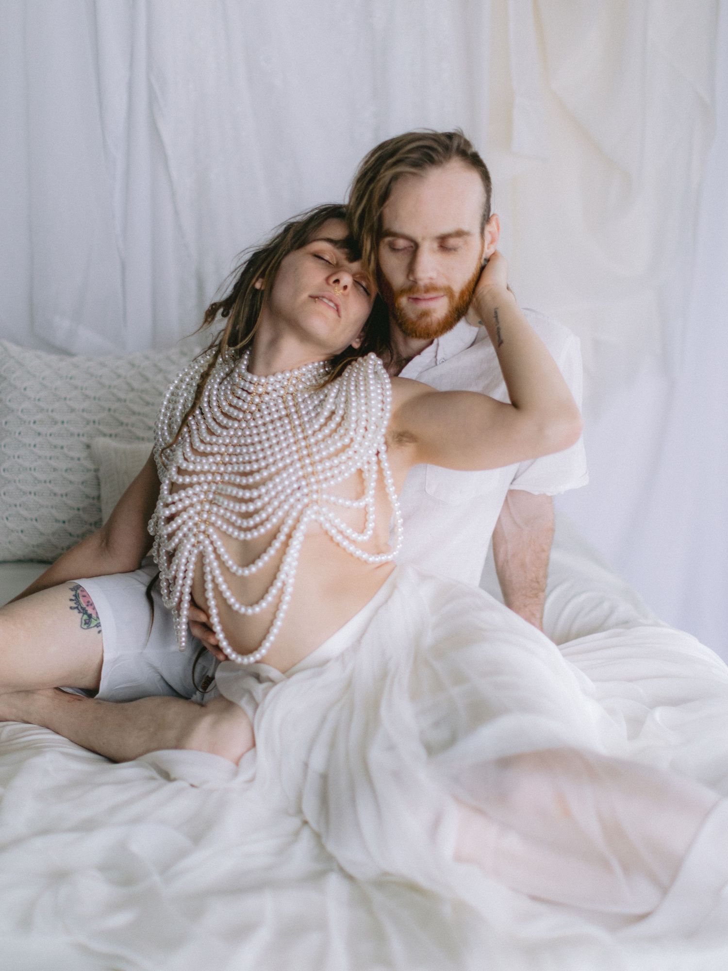 Couple in intimate embrace on white bedding, woman wearing pearl body chain in dreamy studio setting.