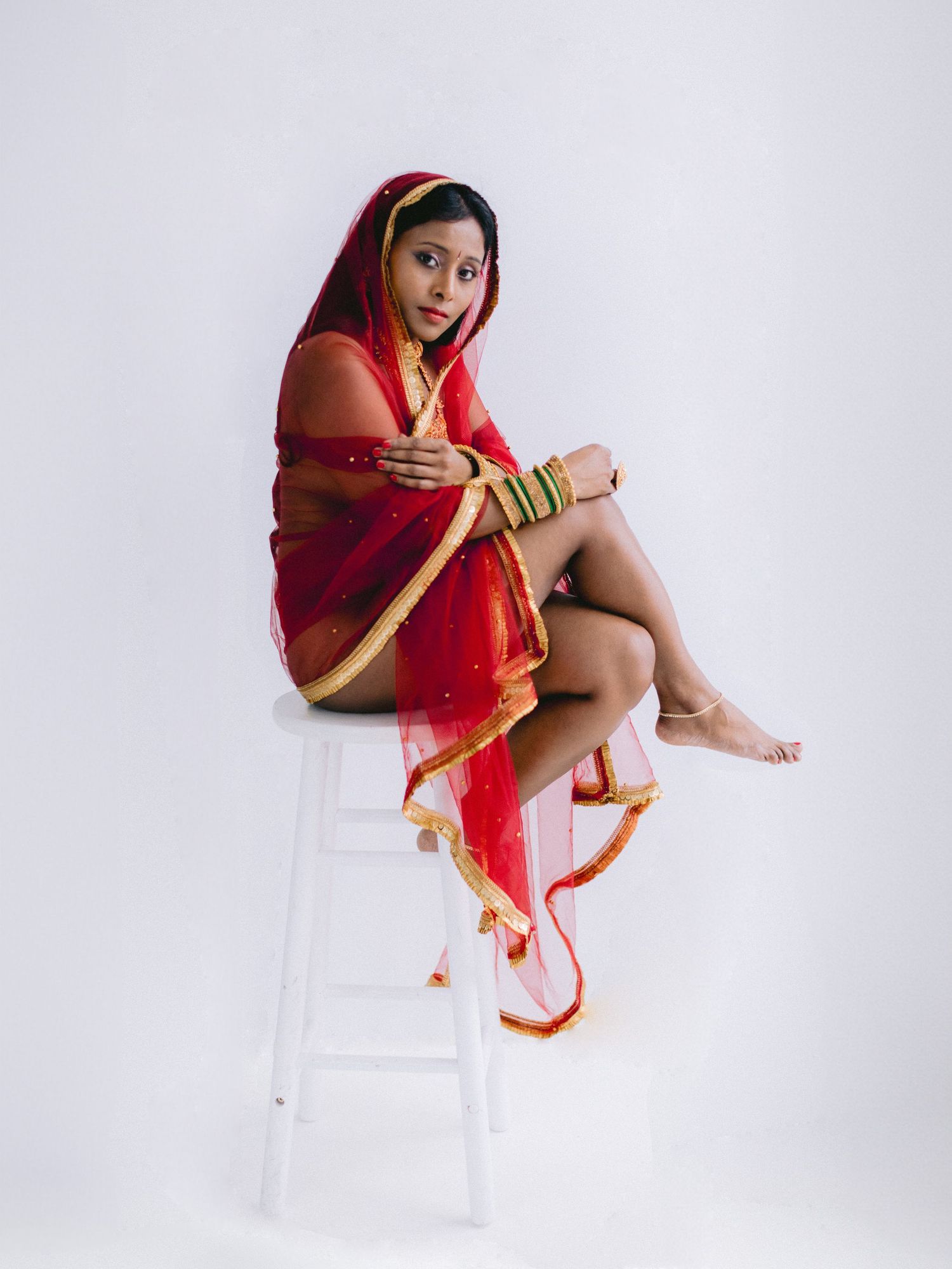 woamn in traditional red sari and bangles poses artistically on white chair against light background.