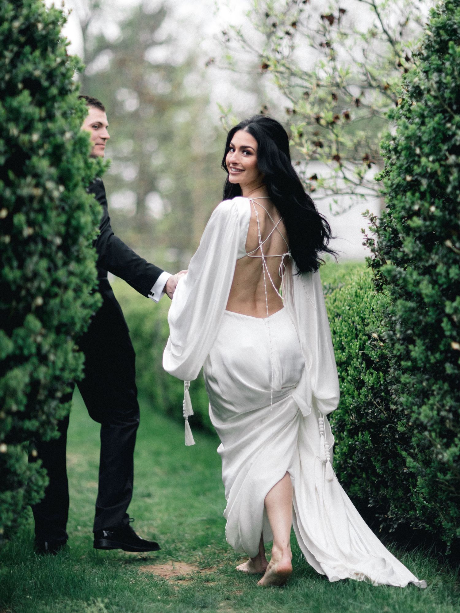 Bride in elegant backless white gown walking through lush garden hedges with groom.
