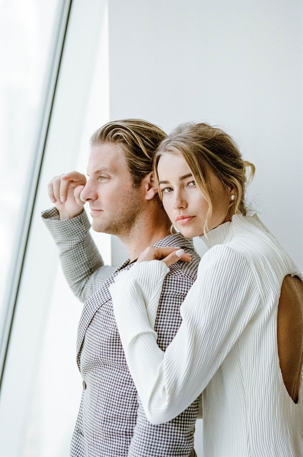 Stylish couple standing by a bright window, woman resting chin on mans shoulder.