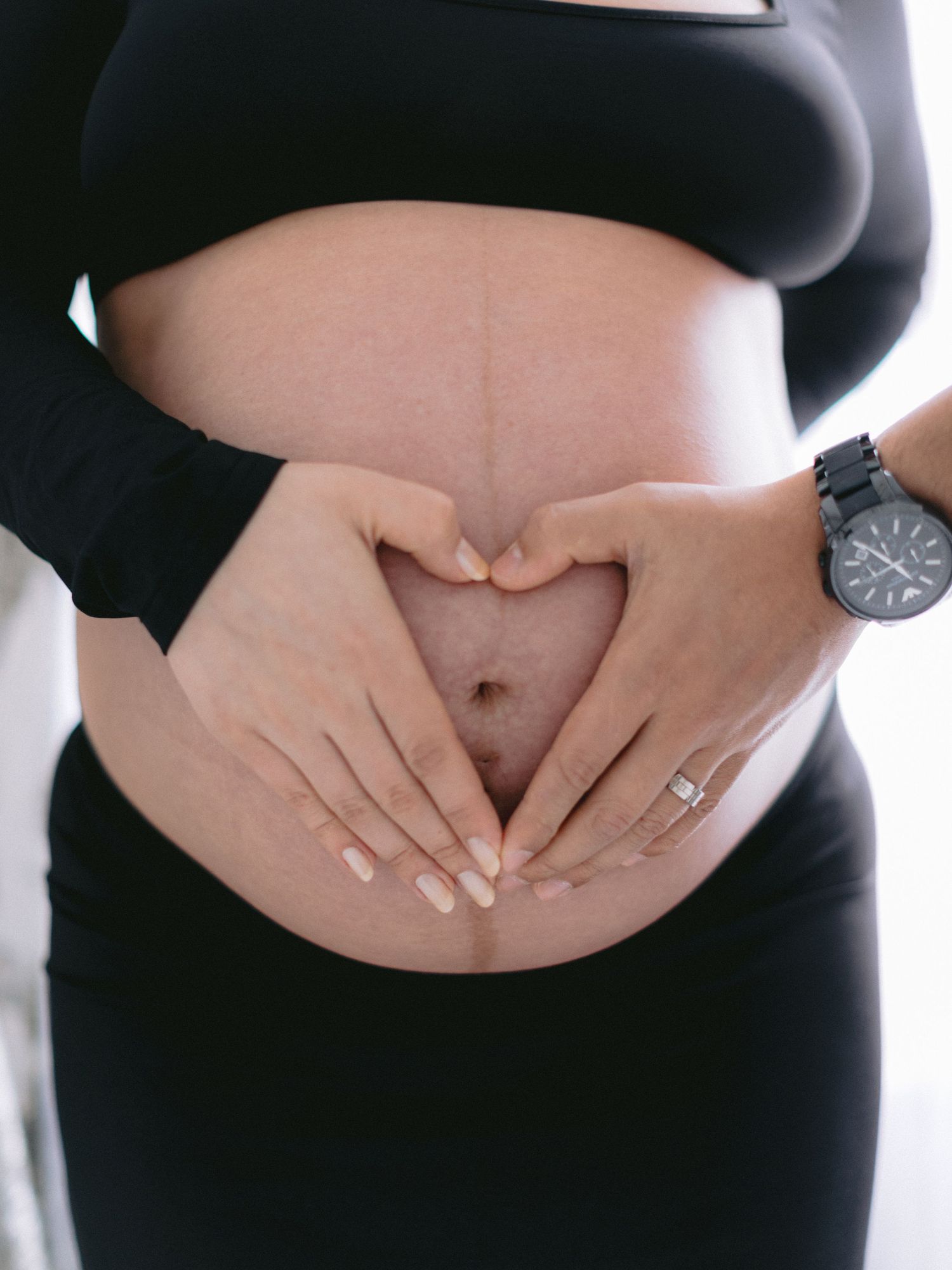 Close-up of pregnant couple forming heart shape with hands on baby bump in black maternity outfit.