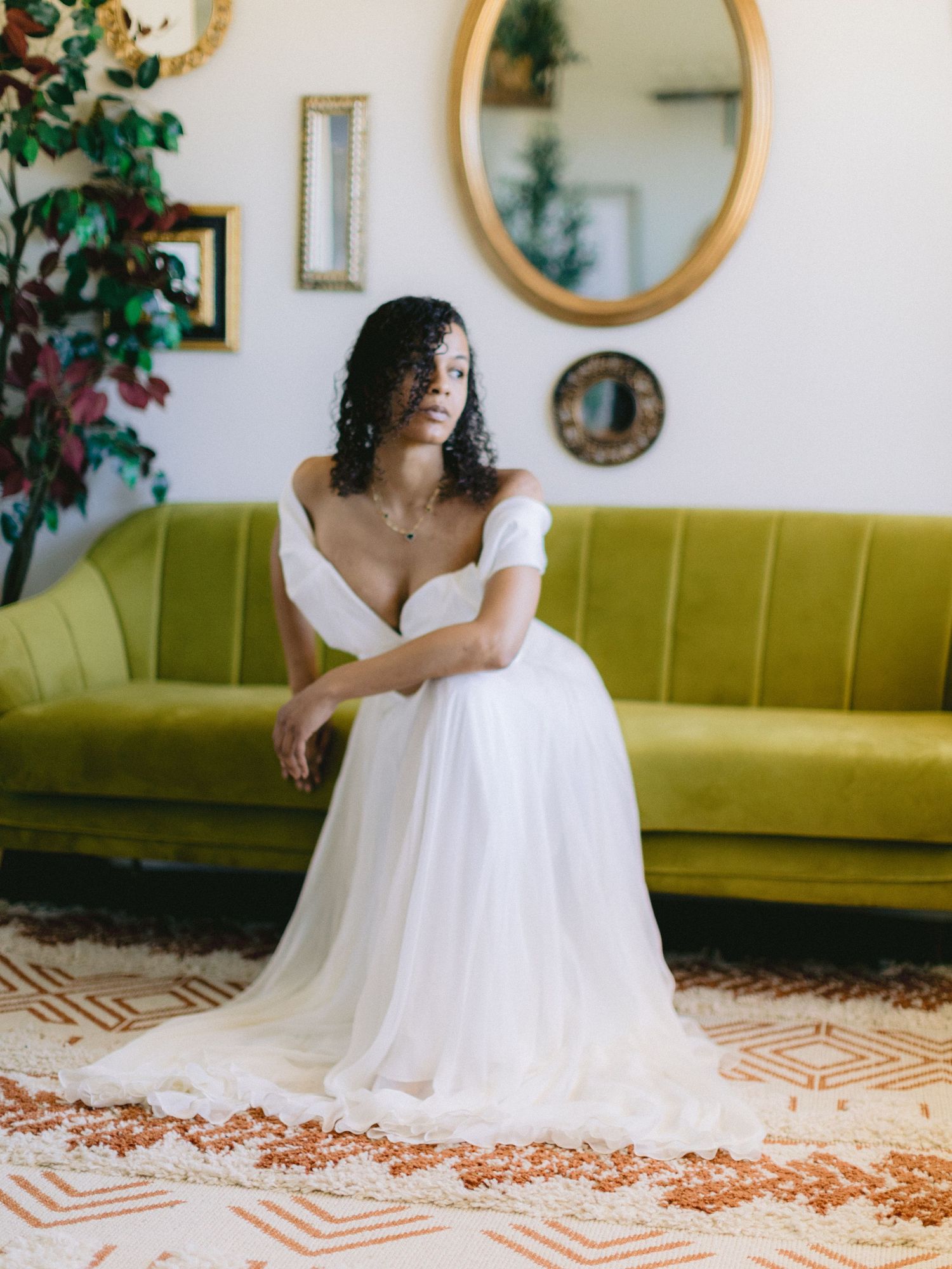 Bride in off-shoulder white tulle gown seated on green velvet sofa in vintage-styled room.