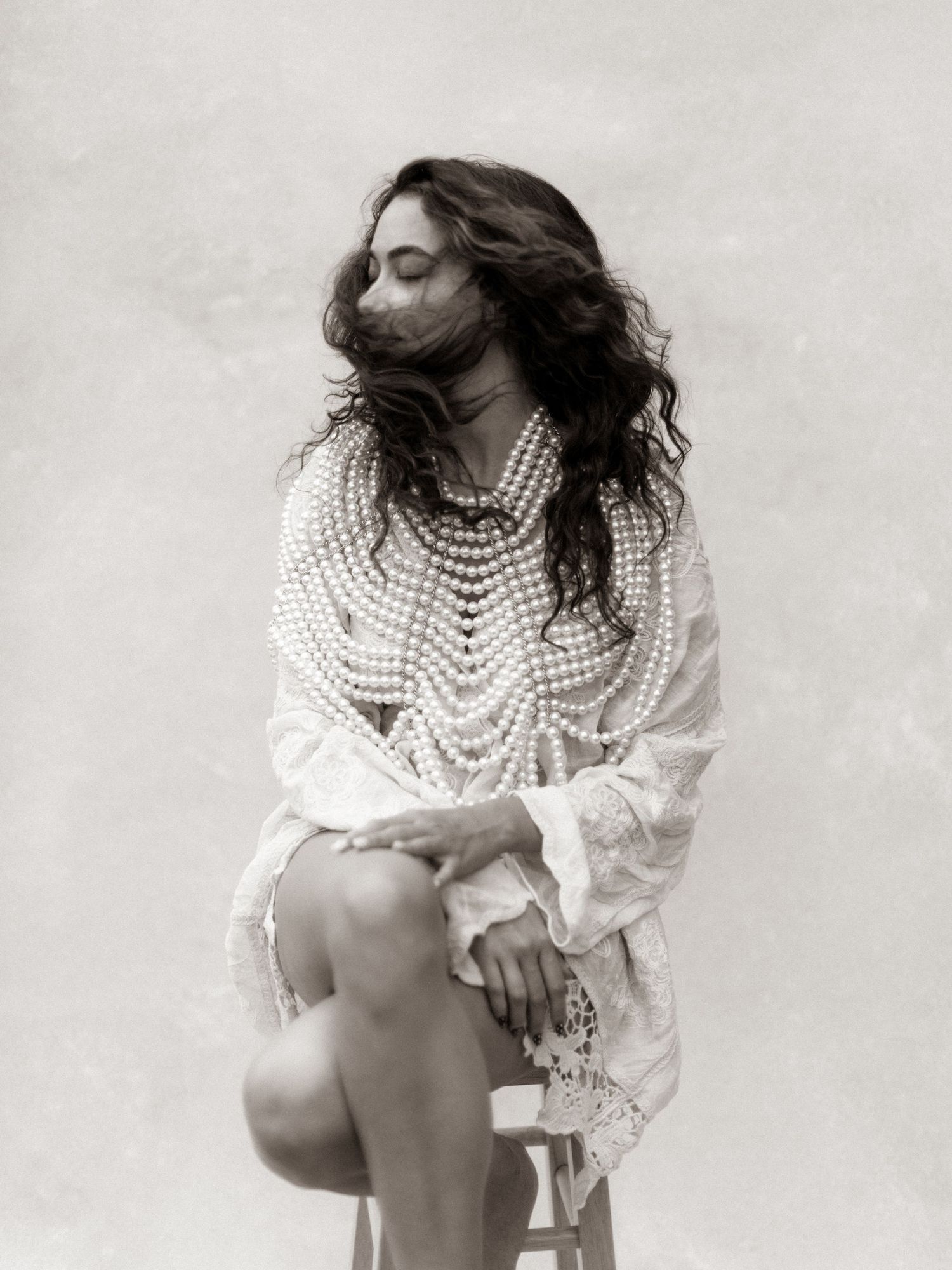 Woman seated in white lace top adorned with layered pearl necklaces in vintage black and white photo.