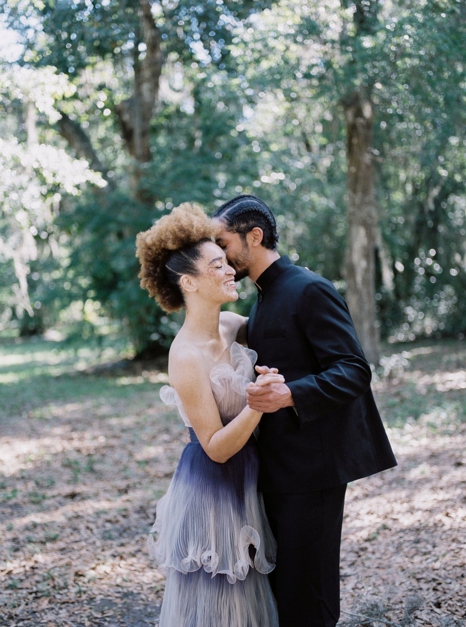 Joyful couple dancing outdoors, woman in gradient blue ruffle gown, man in black suit among trees.