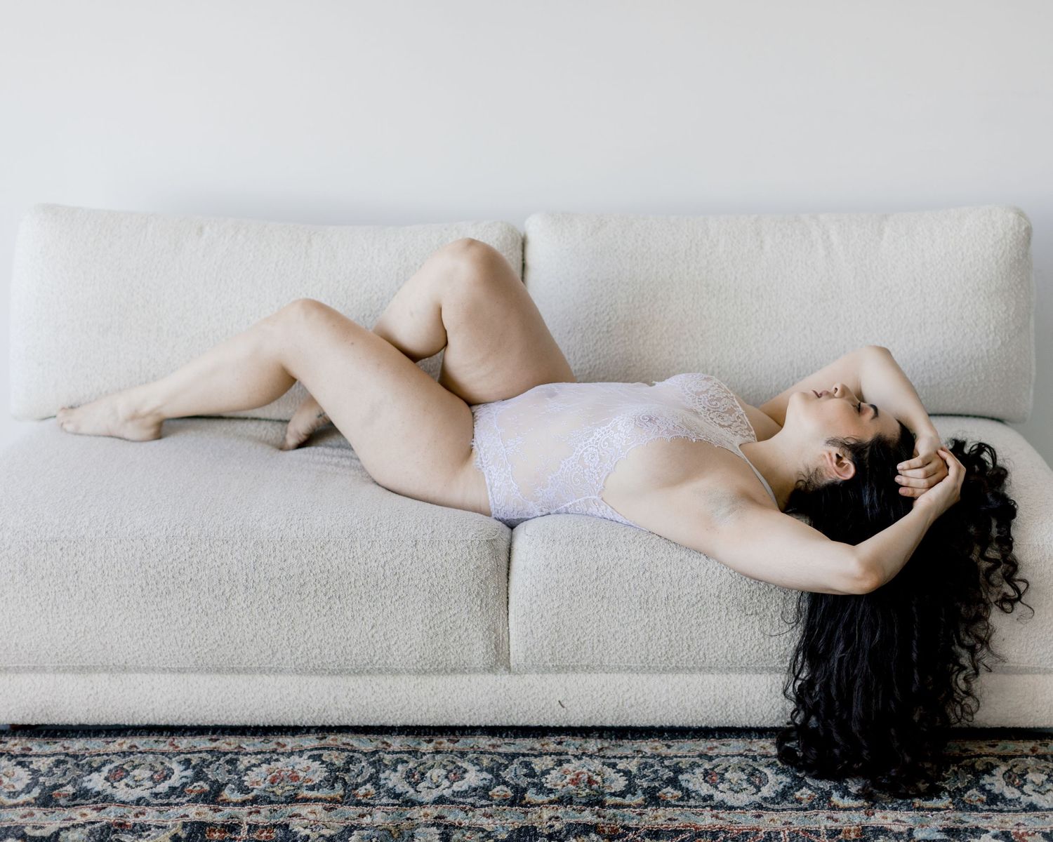 Woman in white lingerie reclining gracefully on a cream boucle sofa over a patterned rug.
