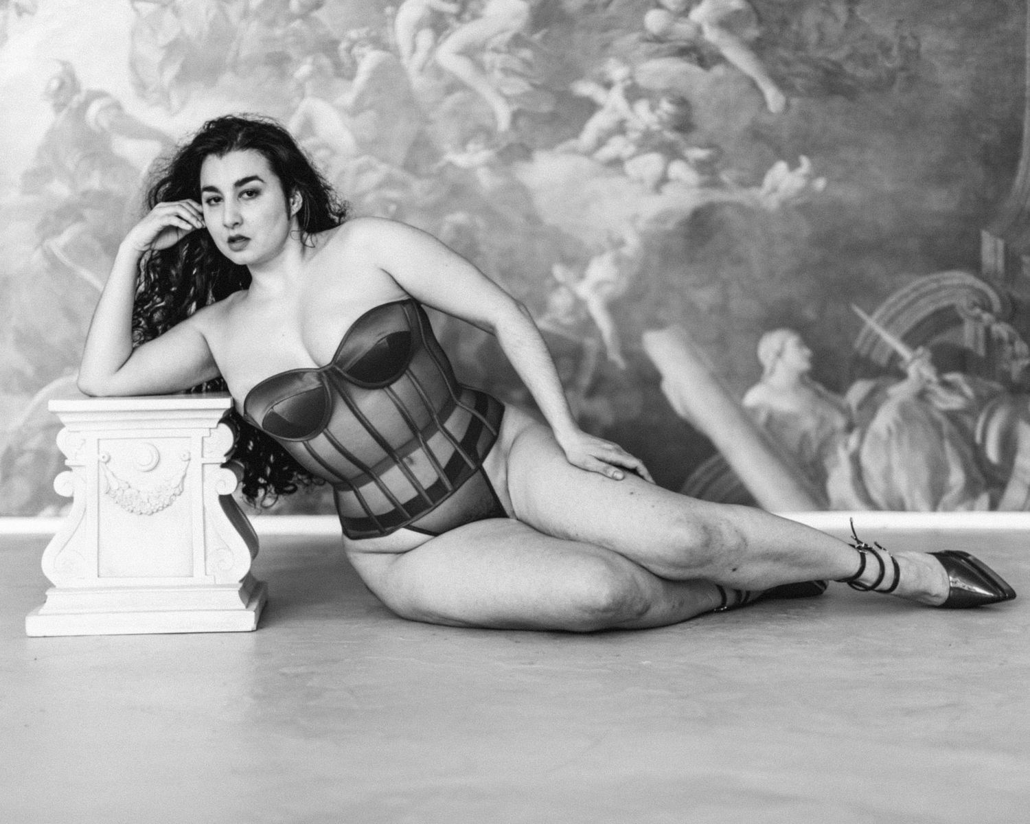 Black and white photo of a woman posing in a corset bodysuit beside a white pedestal with a mural backdrop.