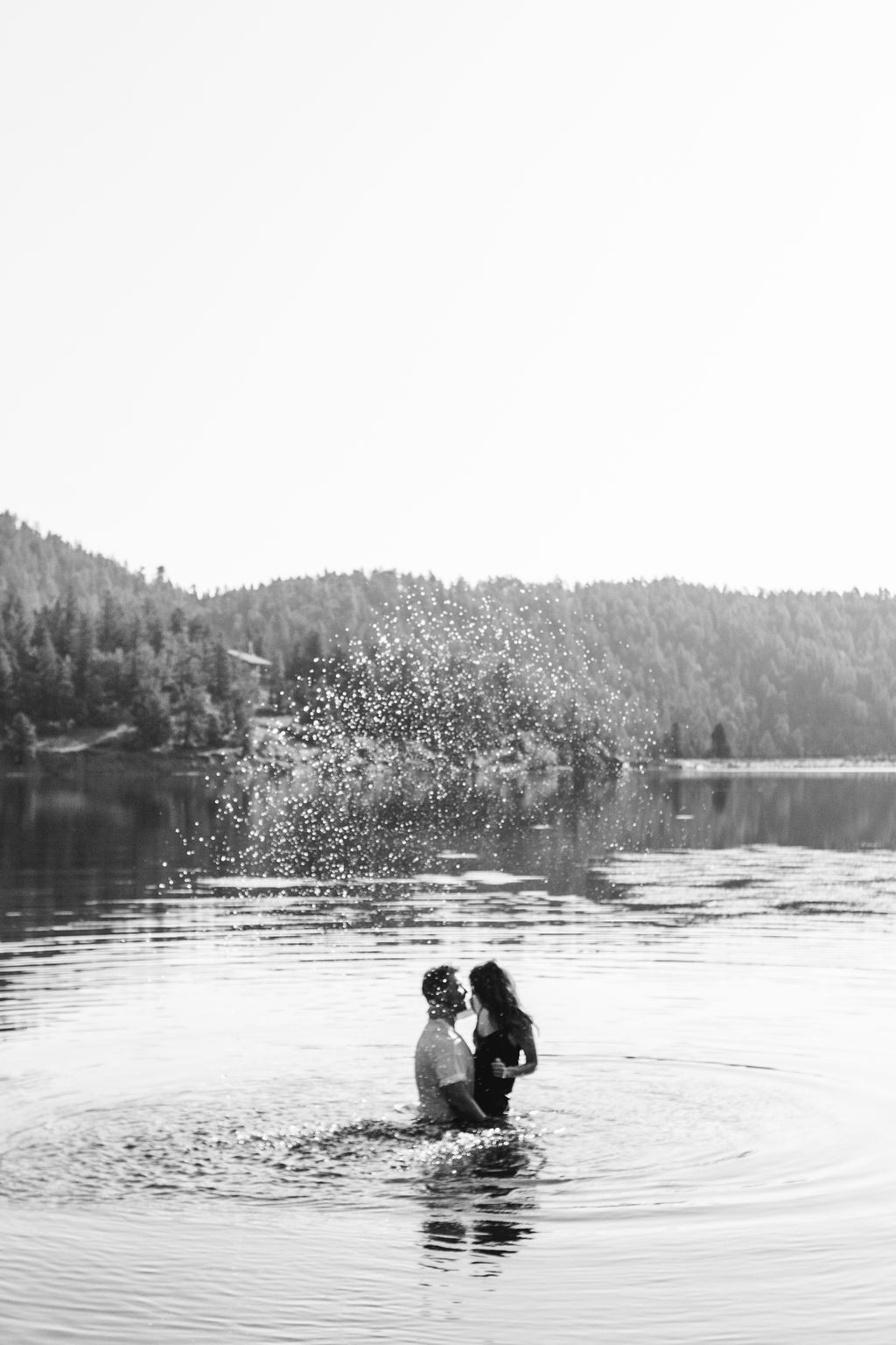 Black and white photo of couple kissing in lake with water splashing and forest backdrop.