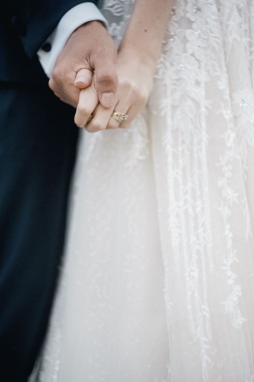 Bride and groom holding hands, showcasing diamond engagement ring against lace wedding dress.