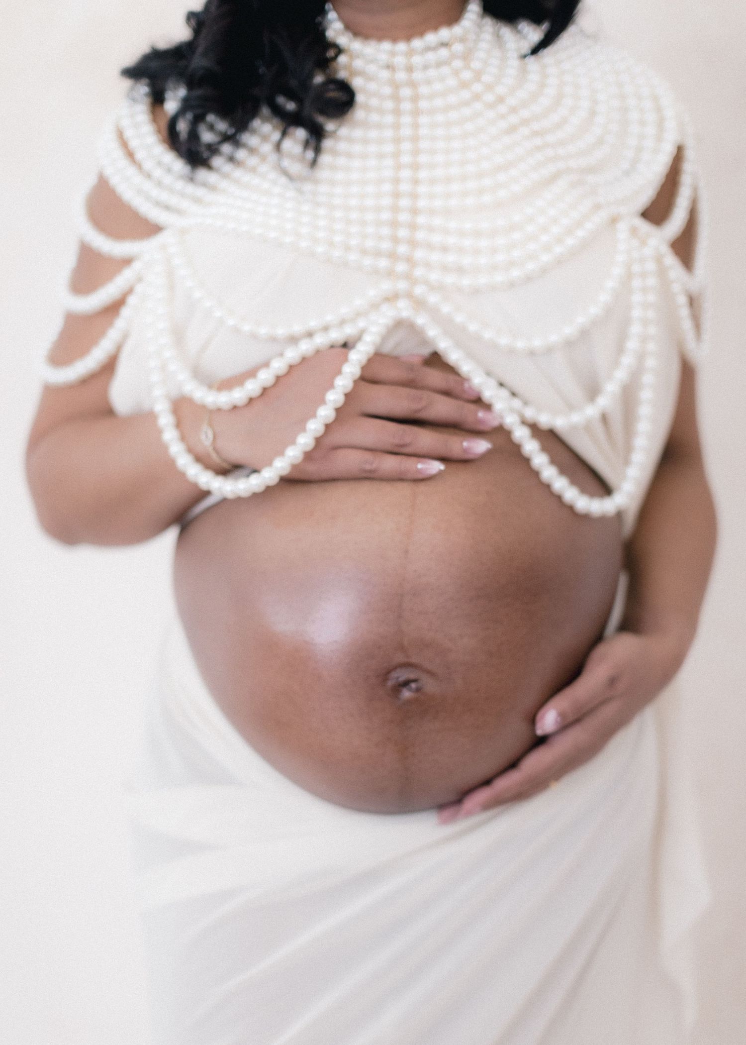 Pregnant woman wearing elegant pearl body jewelry draped over white fabric, cradling her bare baby bump in philadelphia matenity photographe