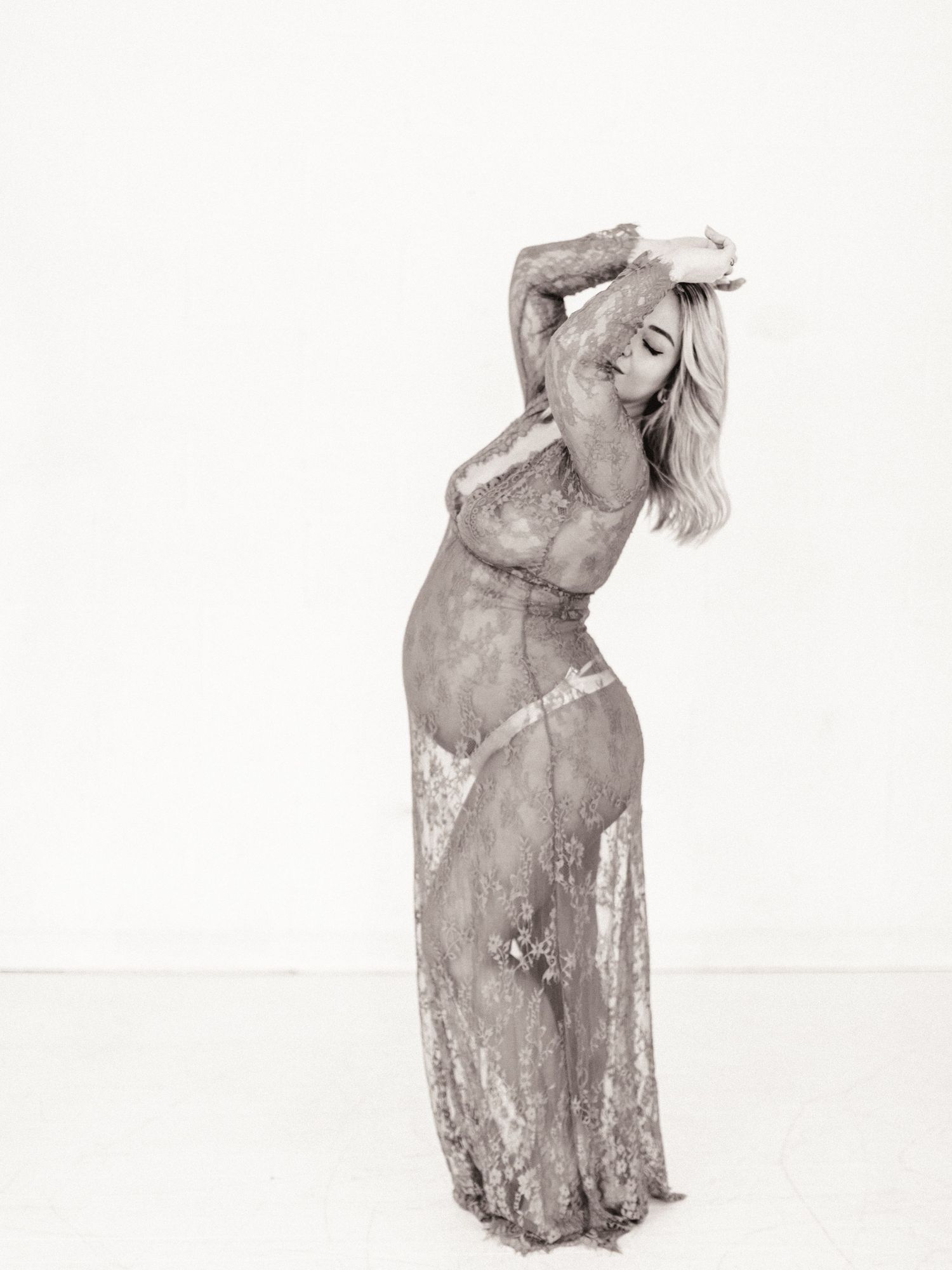 Woman in sheer lace gown poses elegantly with arms raised in a black and white fashion portrait.