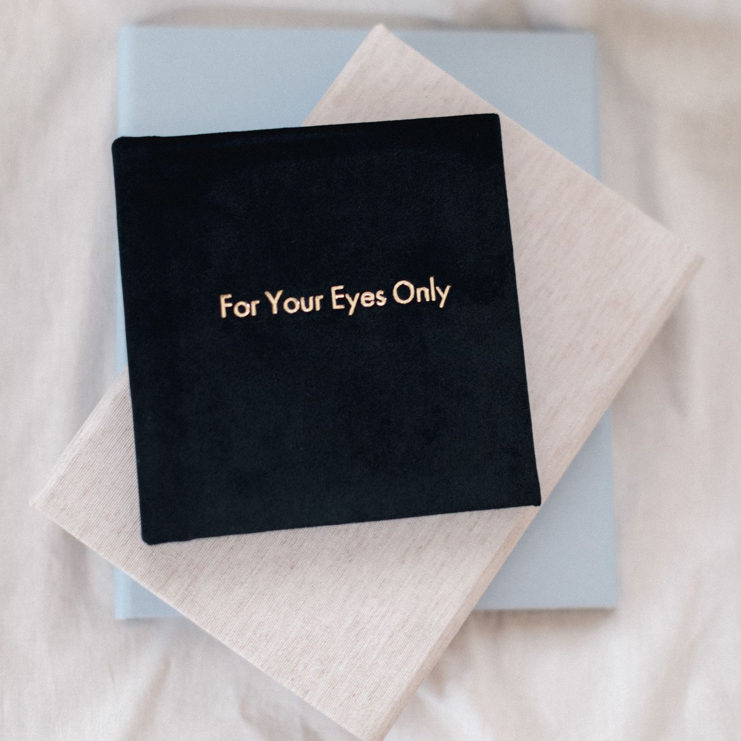 Black velvet album with gold text reading For Your Eyes Only, stacked on beige and blue books on white fabric.