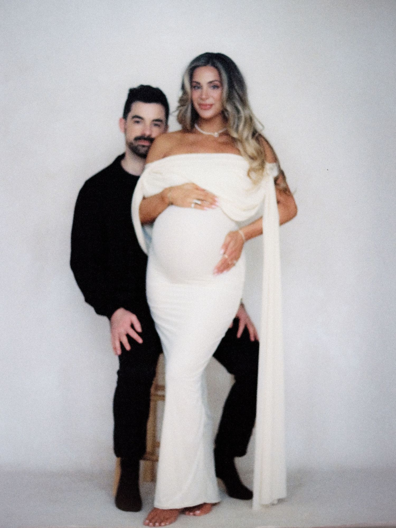 Pregnant woman in elegant white off-shoulder gown poses with partner against white studio background.