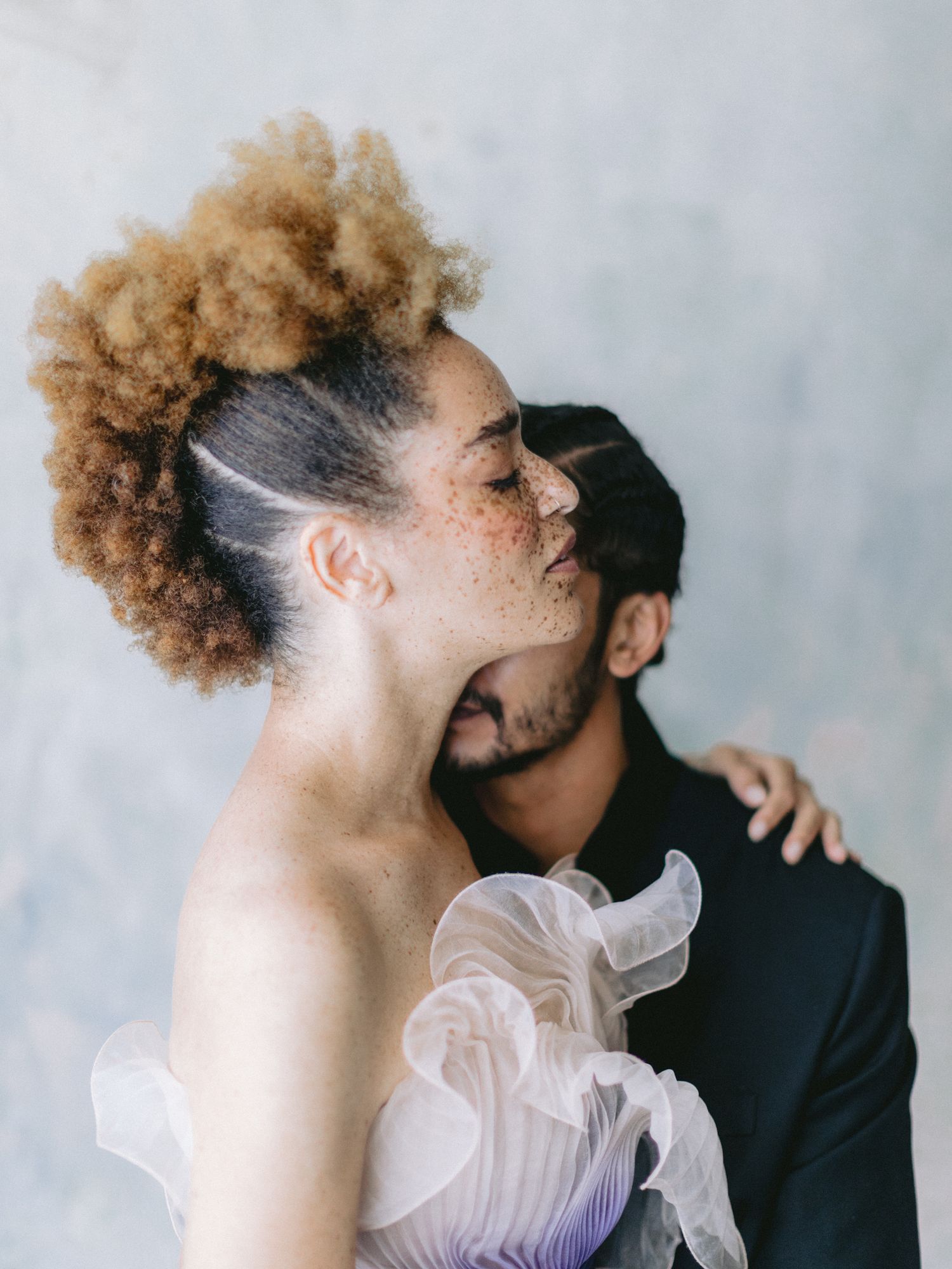 Romantic couple embracing, woman with natural afro updo in strapless ruffle gown kissed by partner.