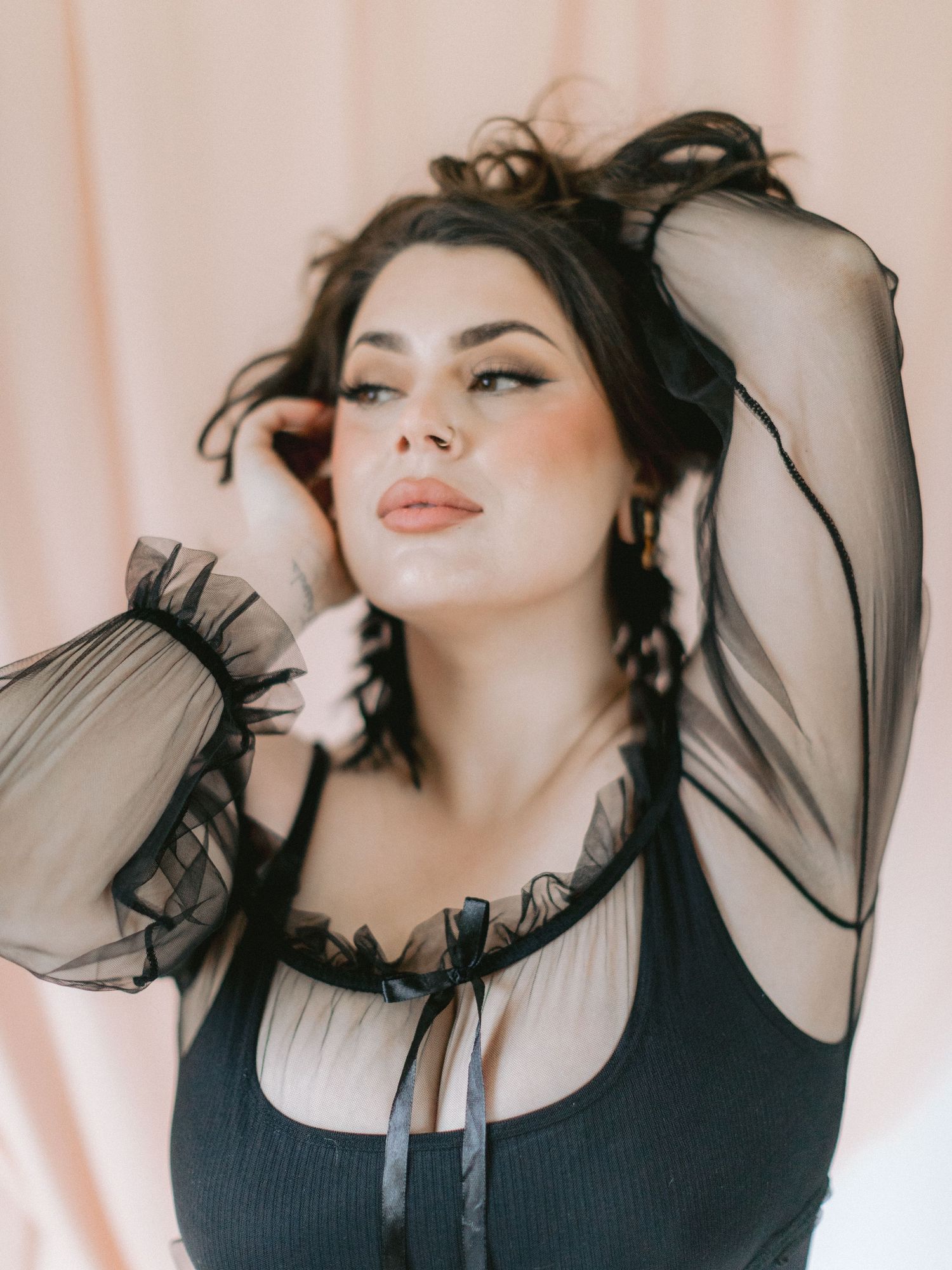 Woman in black sheer-sleeve top poses gracefully with hands in dark hair against a soft pink background.