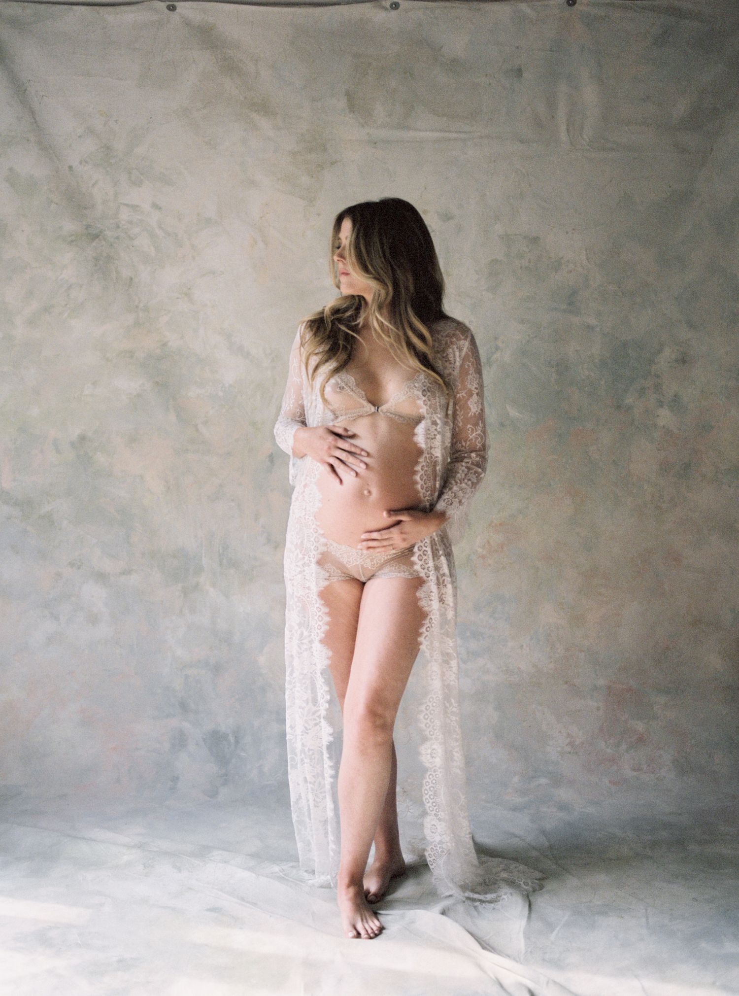 Pregnant woman in sheer white lace robe and lingerie posing in a studio for a maternity photoshoot.
