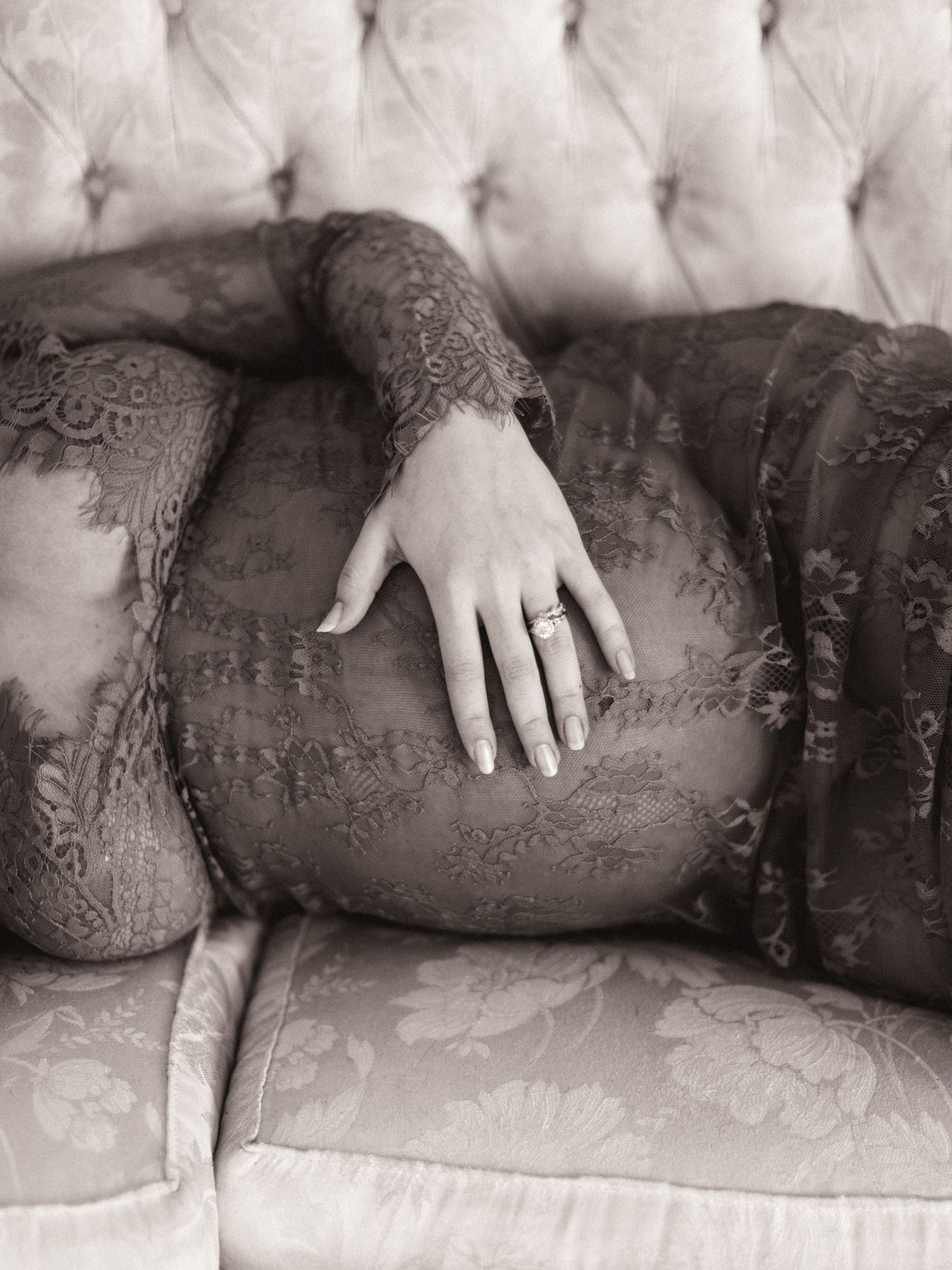 Black and white maternity photo of pregnant woman in lace dress resting on tufted sofa, hand on belly with diamond ring.