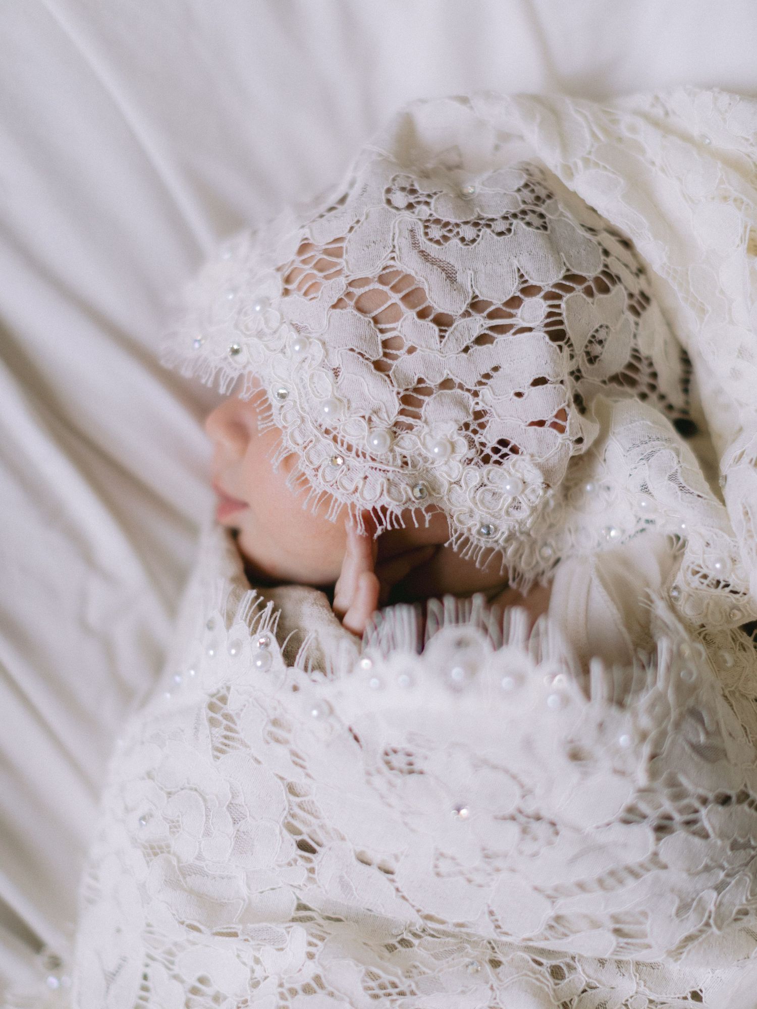 A newborn baby wrapped in delicate white lace fabric and bonnet, peacefully sleeping in soft white surroundings.