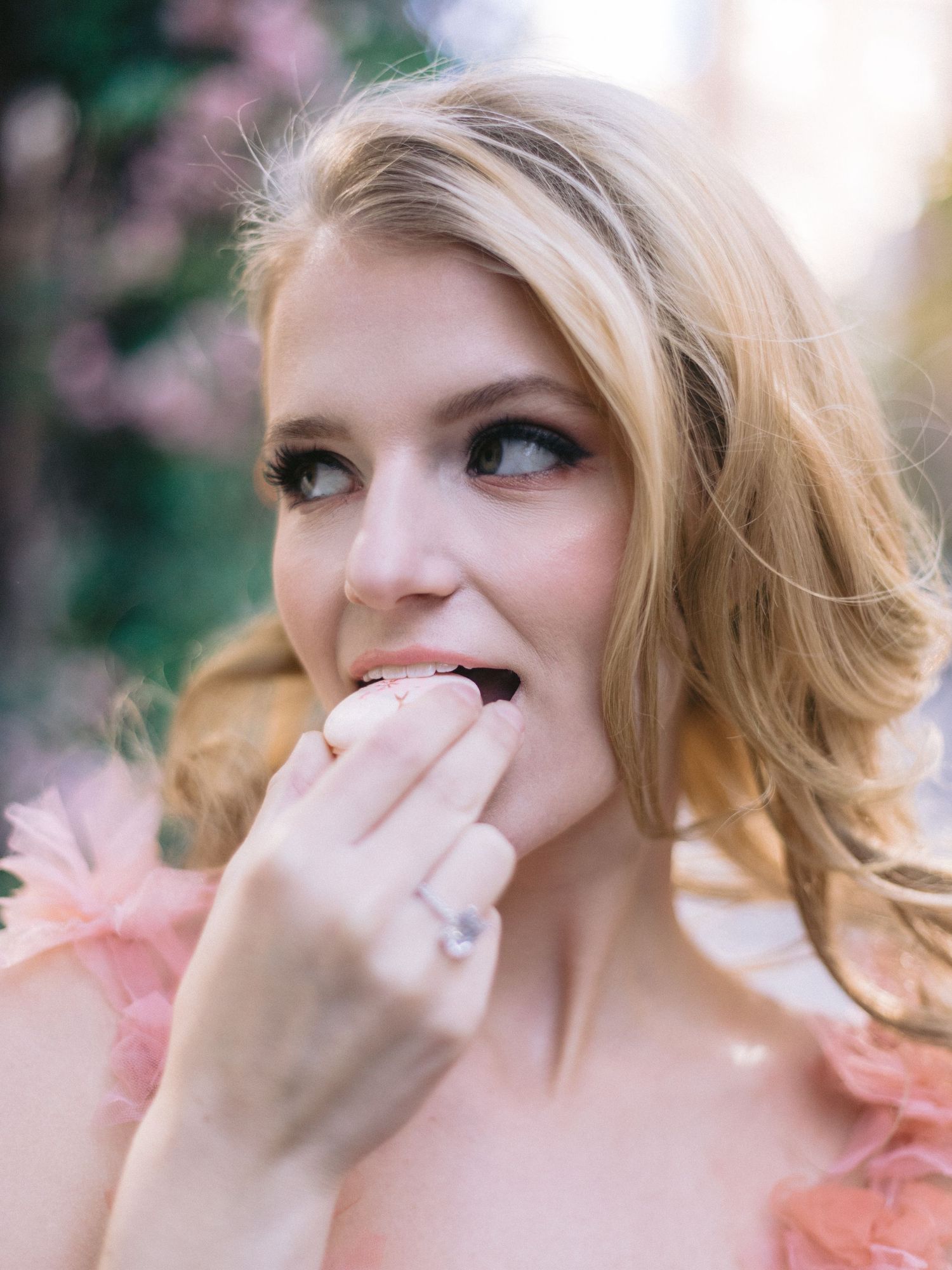 Blonde bride in pink ruffled dress eating macaron, displaying diamond engagement ring outdoors.