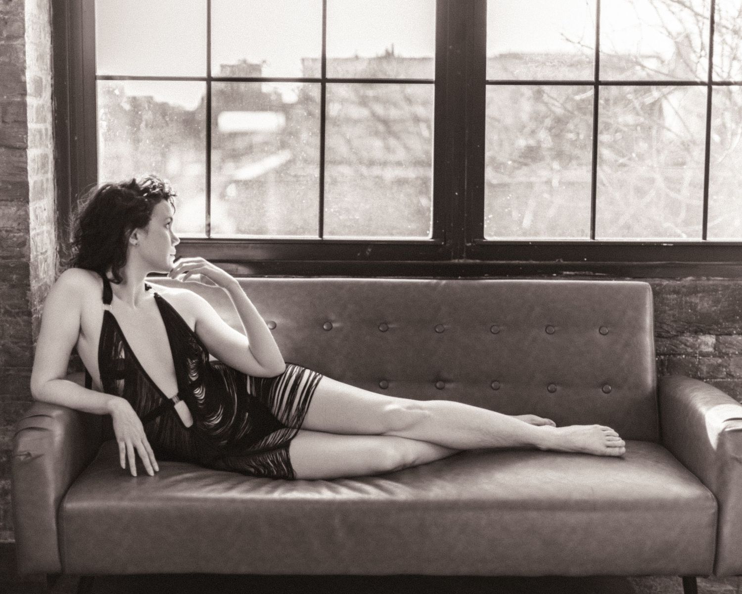 Black and white photo of a woman in a sequined dress reclining gracefully on a leather sofa near large windows.