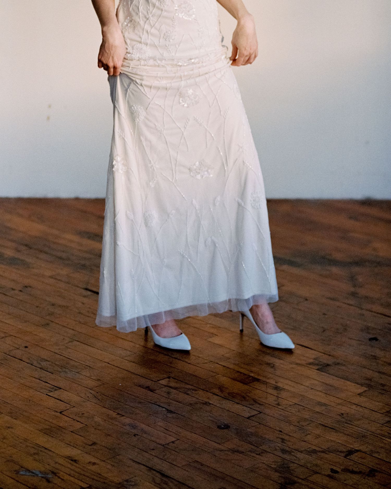 Woman in embroidered white bridal gown with blue heels standing on rustic hardwood floor.