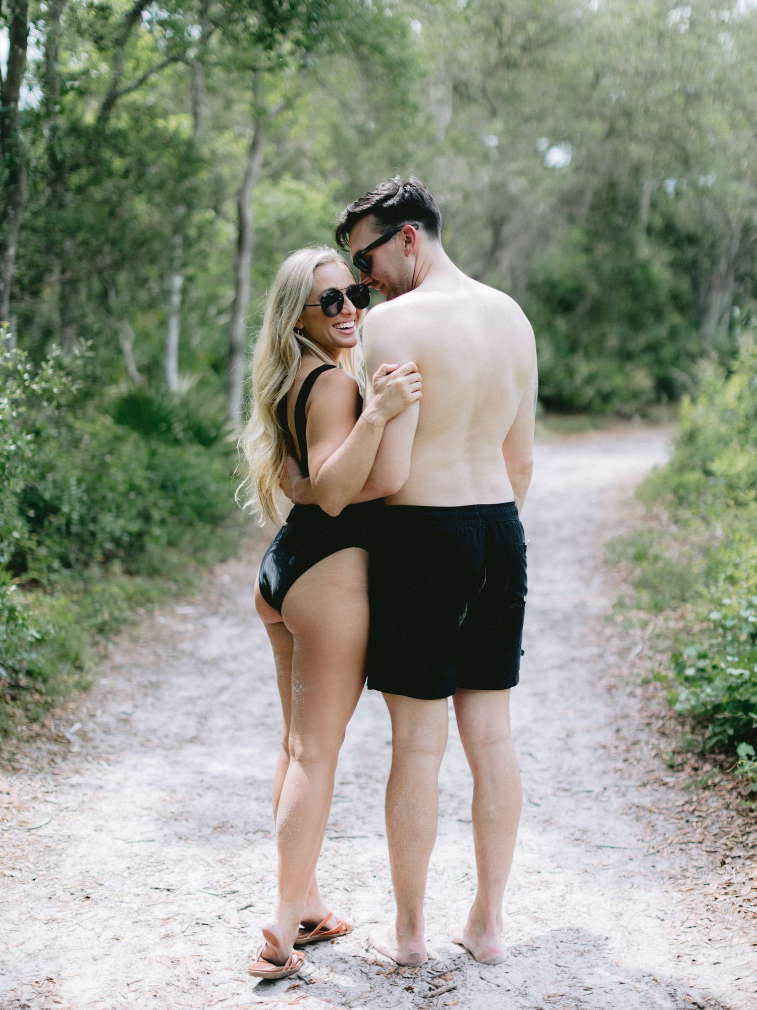 Couple in swimwear embracing on sandy forest path during outdoor engagement photoshoot.