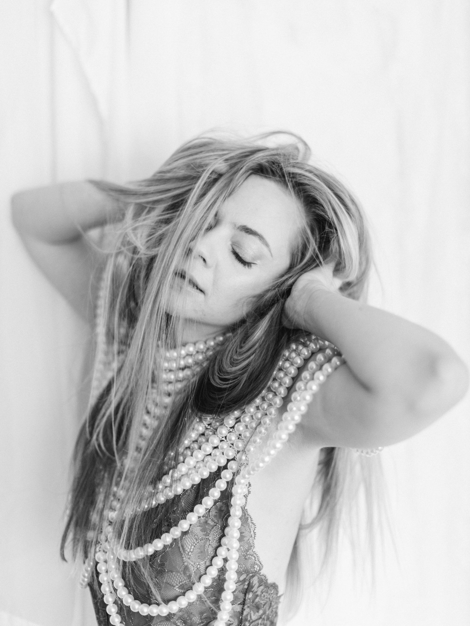 Black and white photo of a woman with long hair wearing layered pearl necklaces, posing gracefully with eyes closed.