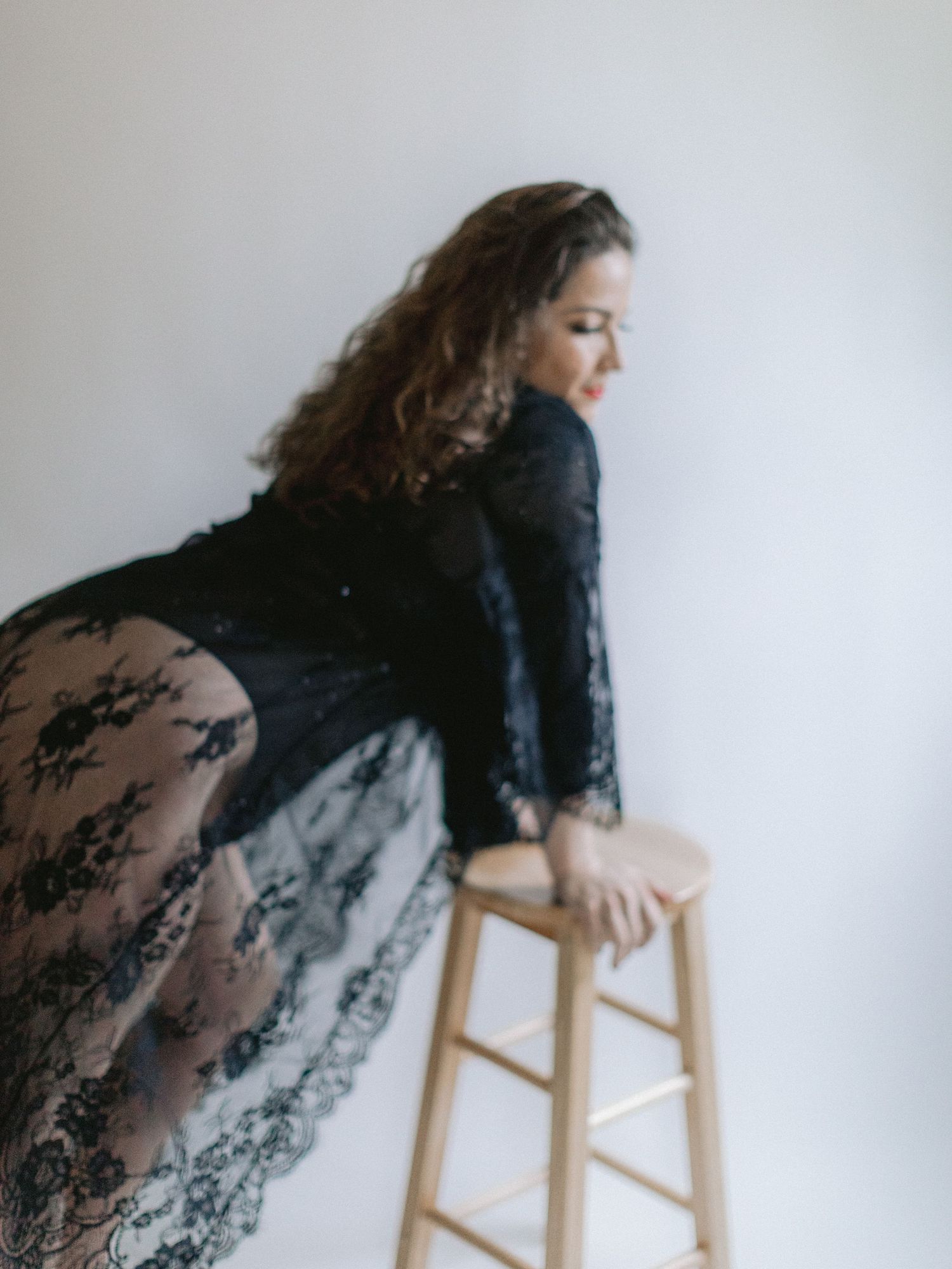 A woman in a black lace dress poses artistically while leaning against a wooden stool in a minimalist setting.