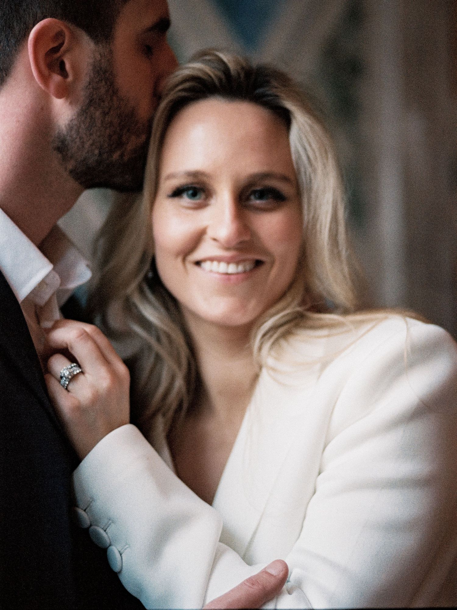 Smiling blonde bride in white blazer with groom kissing her cheek tenderly.