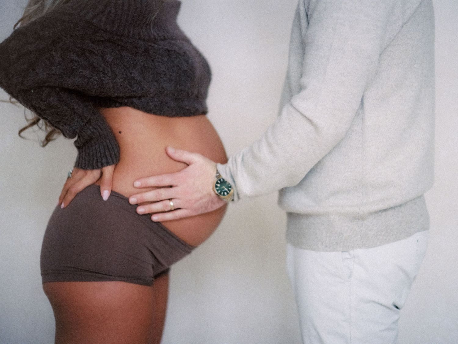 Pregnant woman in brown shorts with partner's hand on baby bump, maternity photo shoot.